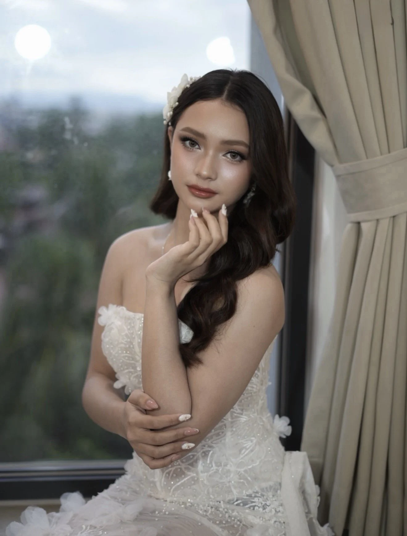 A woman with long dark hair, wearing a strapless white lace dress with floral embroidery, sitting by a window with beige curtains, looking at the camera with a gentle expression.