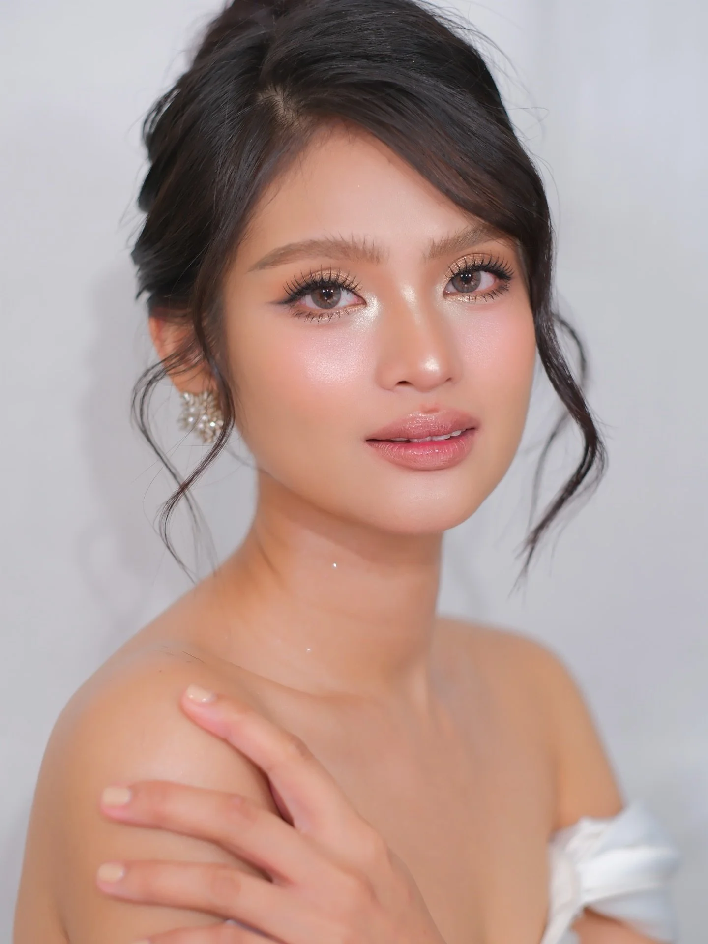 A portrait of a young woman with dark hair styled in loose waves, wearing makeup with a focus on her eyes and lips, and earrings, against a plain background.