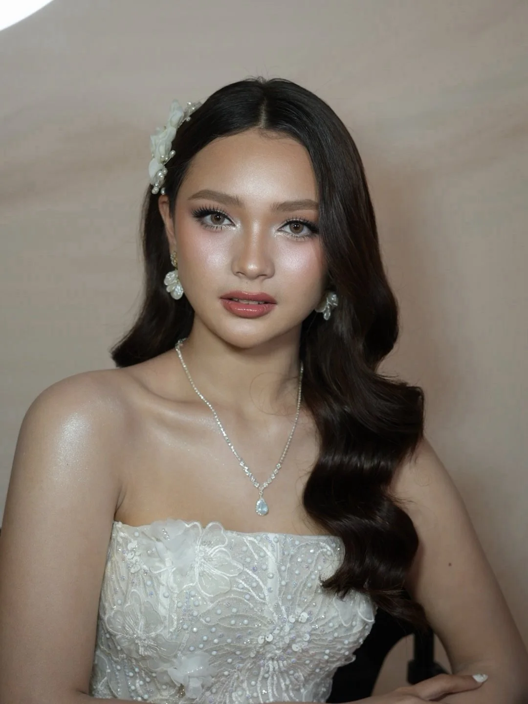 A young woman with long dark hair wearing a white strapless dress with floral embroidery, pearl earrings, a pearl necklace with a pendant, and a white flower hair clip, posing against a neutral background.