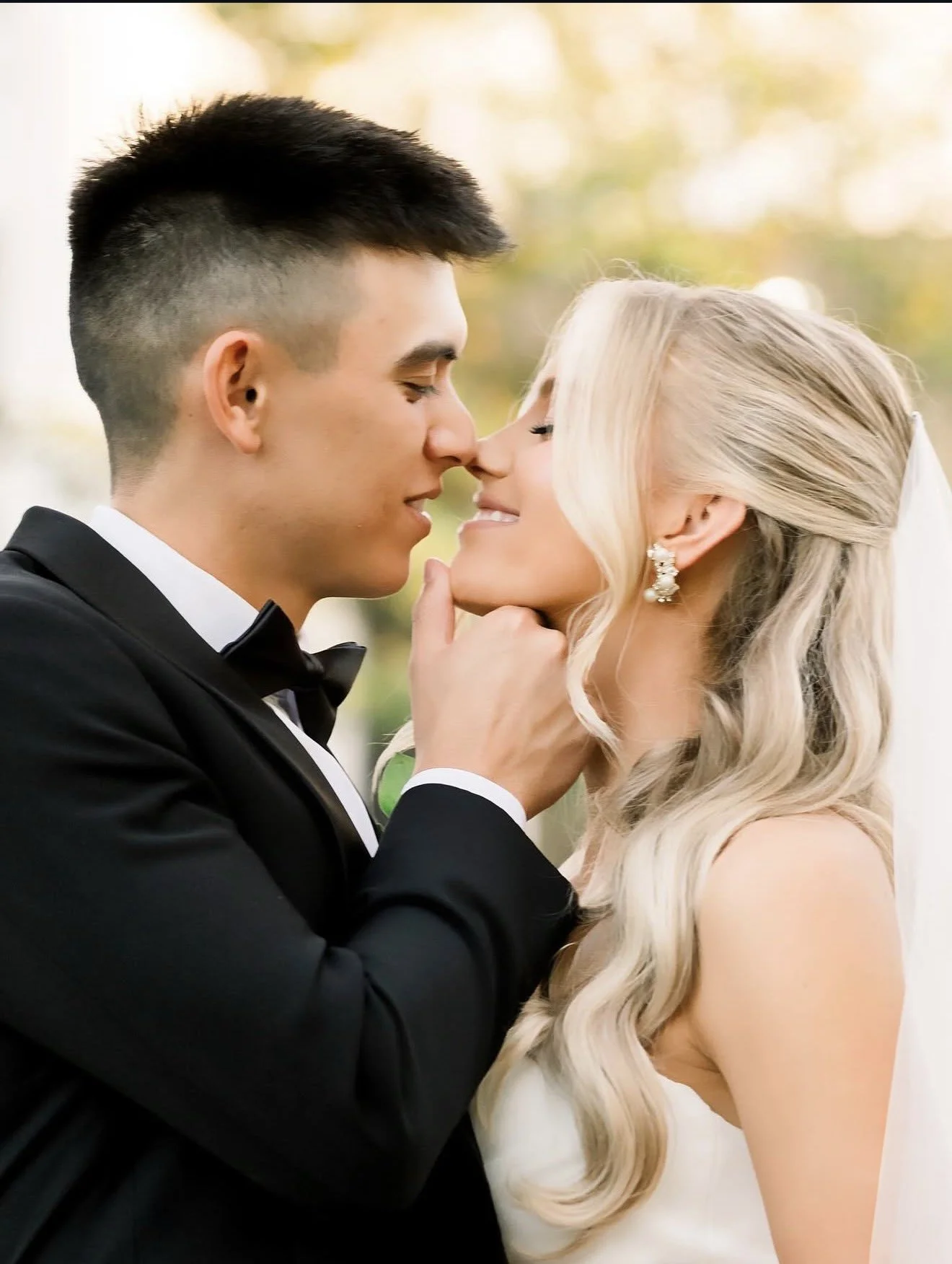 A close-up of a bride and groom touching noses, with their eyes closed, in an outdoor setting with blurred trees in the background.