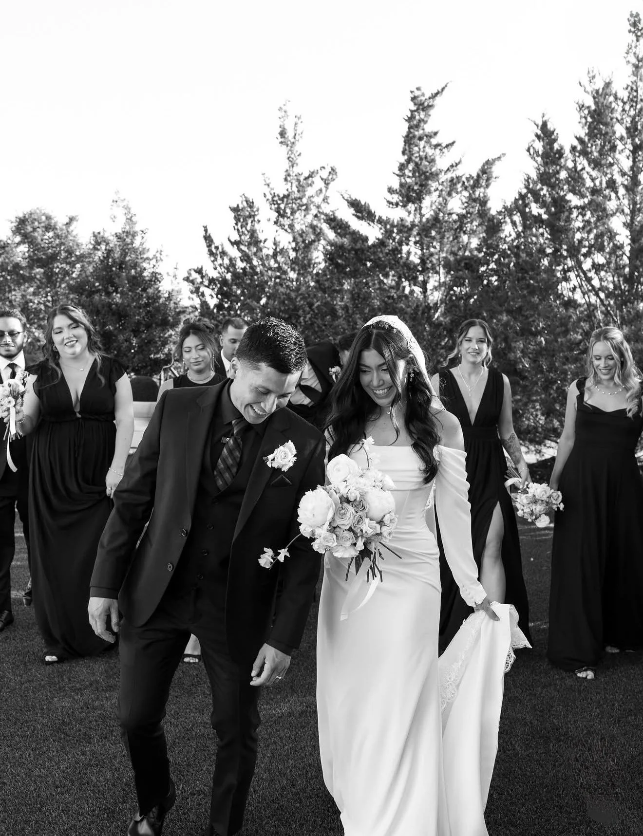 A wedding scene with a bride and groom walking outdoors, surrounded by bridesmaids and groomsmen, all smiling and holding flowers, with trees in the background.