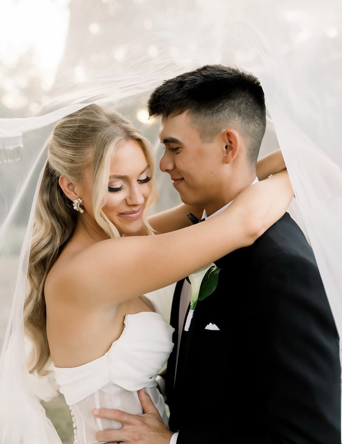 Bride and groom sharing an intimate moment, with the bride wearing a white wedding dress and the groom in a black tuxedo, under a sheer veil.