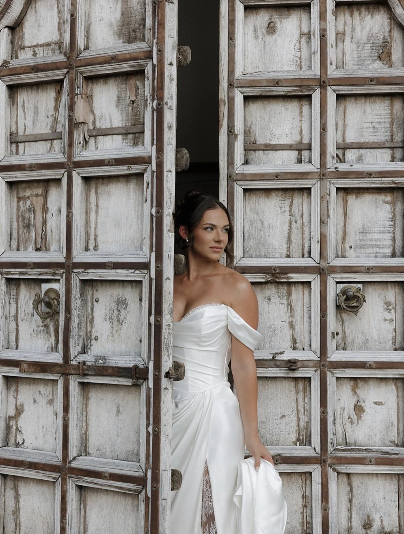 A woman in a white strapless wedding dress stands partially behind a rustic, weathered wooden door, gazing to her left.