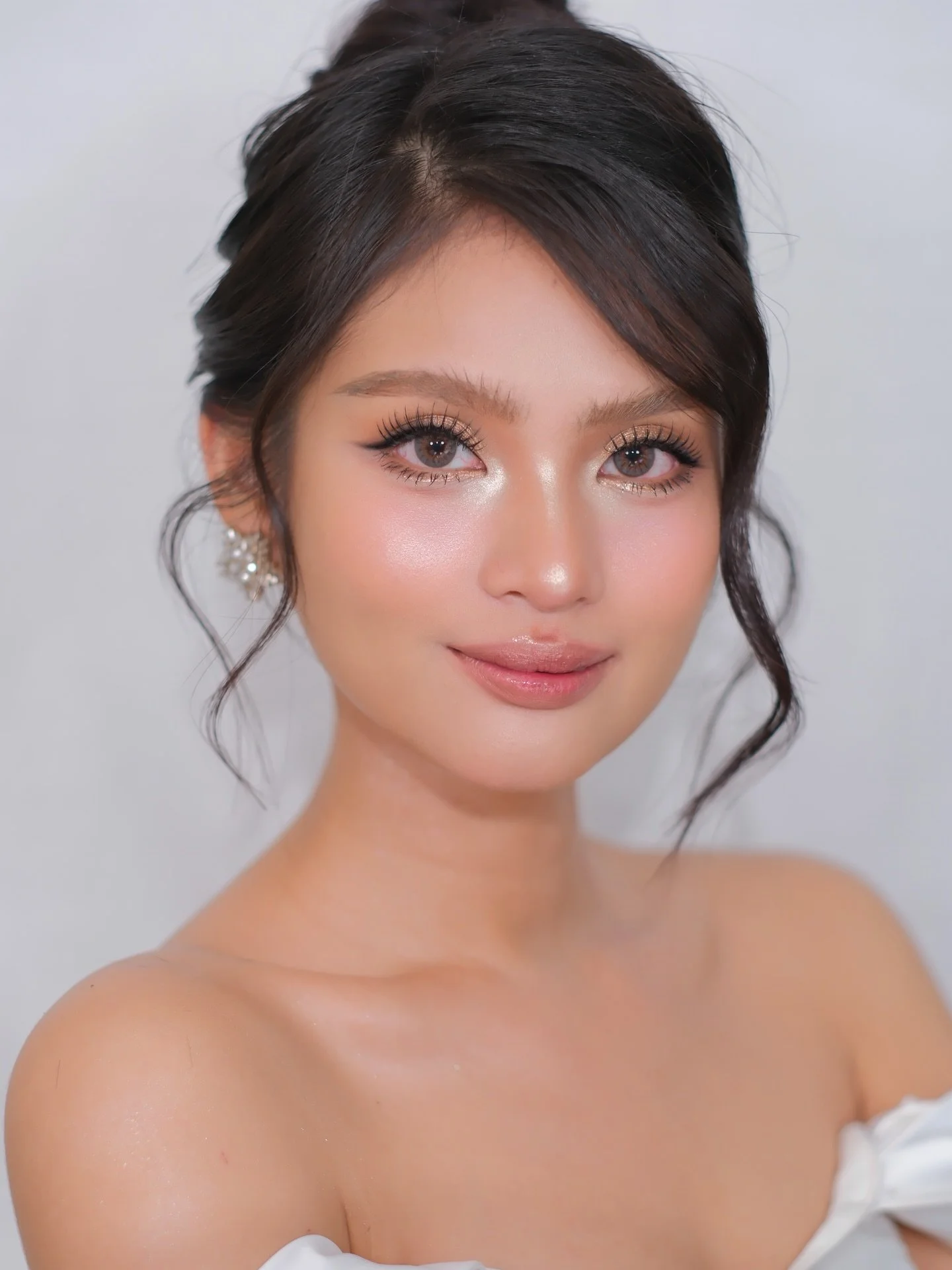 Close-up portrait of a woman with dark hair styled in soft waves, wearing makeup with subtle eyeshadow and glossy lips, and an off-shoulder white top, against a plain light background.