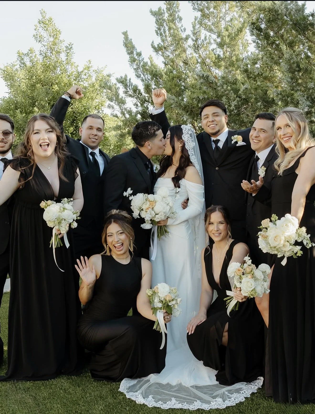 A group of people at a wedding, including the bride and groom kissing in the center, surrounded by bridesmaids and groomsmen, celebrating outdoors with trees in the background.