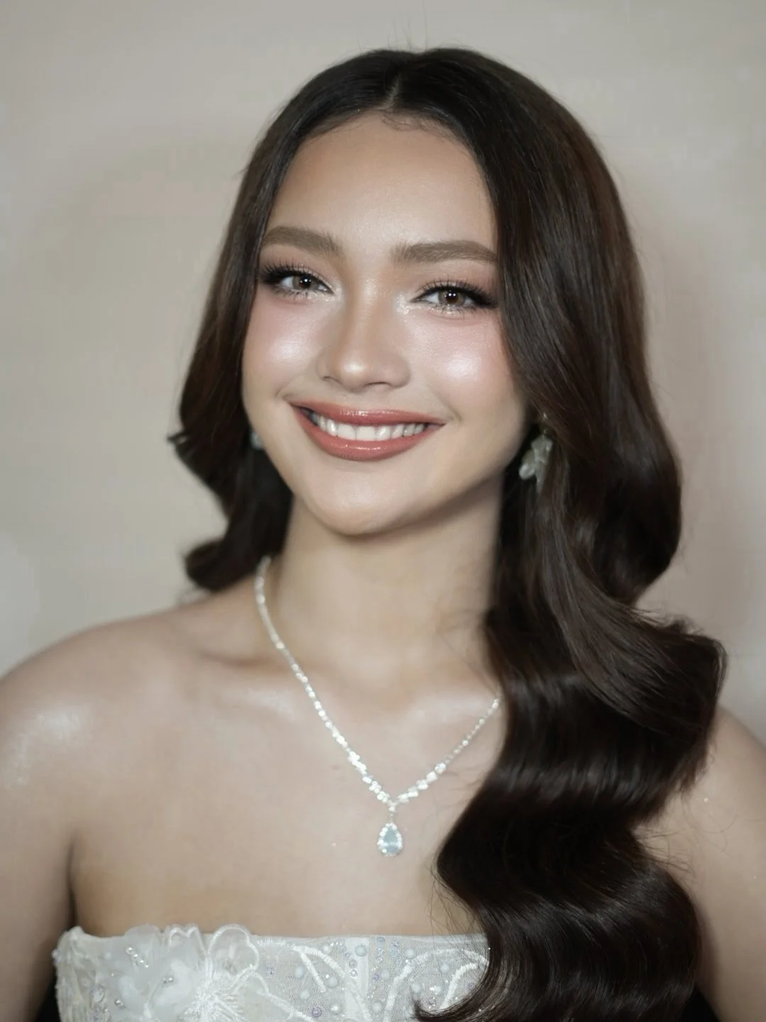 Portrait of a young woman with wavy brown hair, smiling, wearing a white strapless dress and a silver necklace with a teardrop pendant.