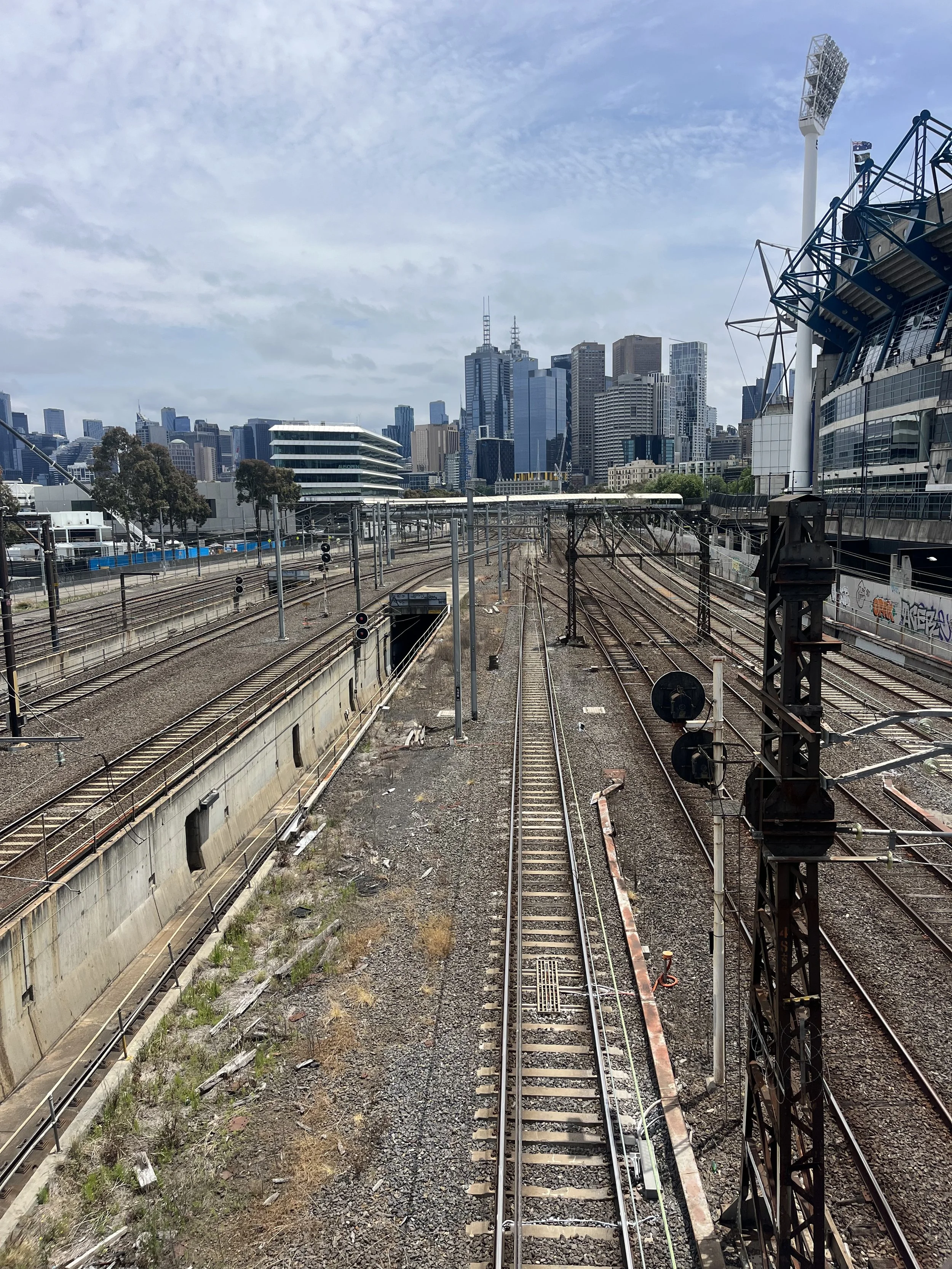 View of train tracks in a city with modern skyscrapers in the background on a partly cloudy day.