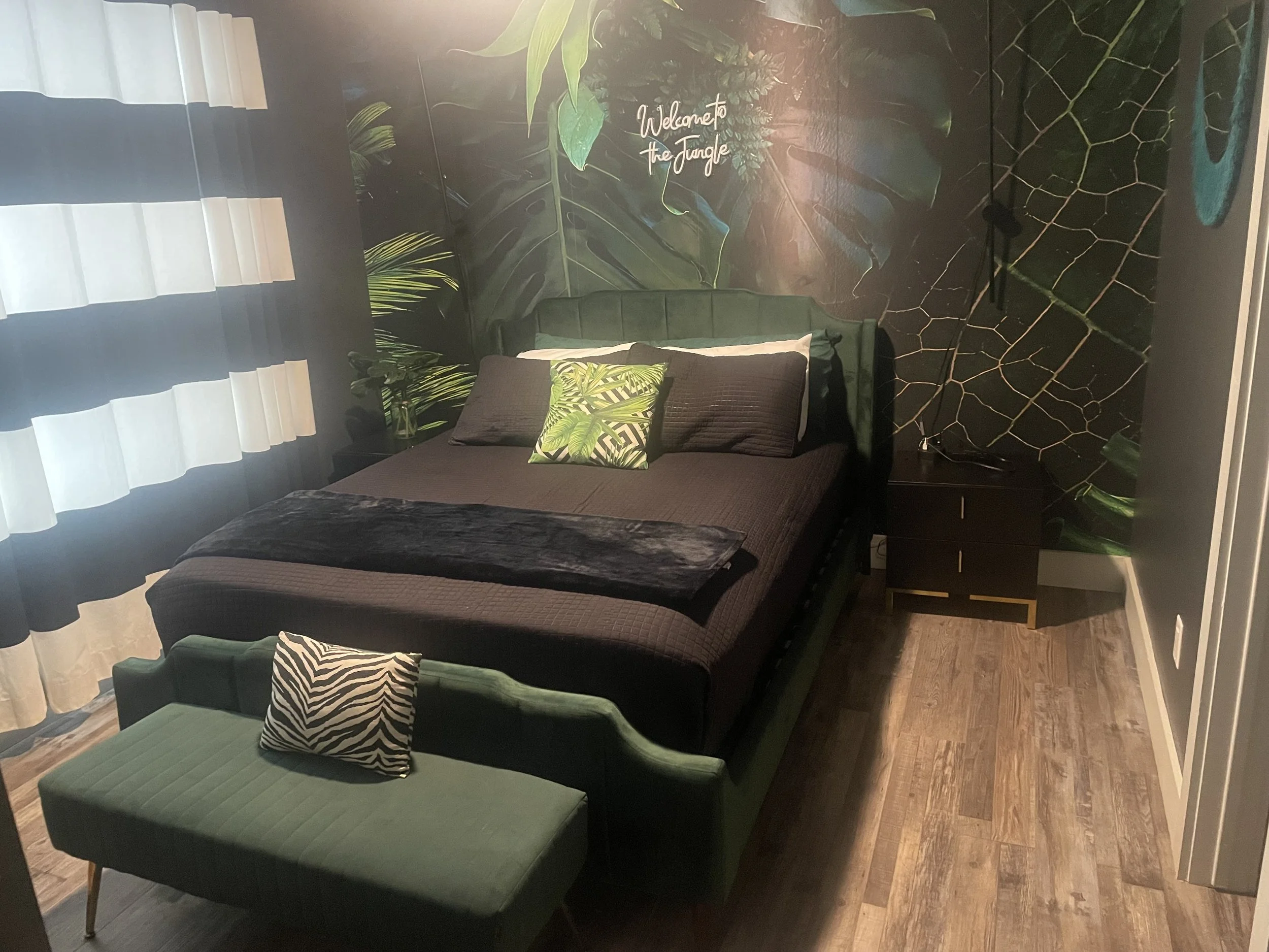 Modern bedroom with a jungle-themed wall mural behind the bed, featuring large green leaves and a quote that says 'Welcome to the Jungle'. The bed has black bedding and decorative pillows, including one with a tropical leaf pattern. There are black nightstands and a small green bench at the foot of the bed. A window with white and black curtains lets in natural light.