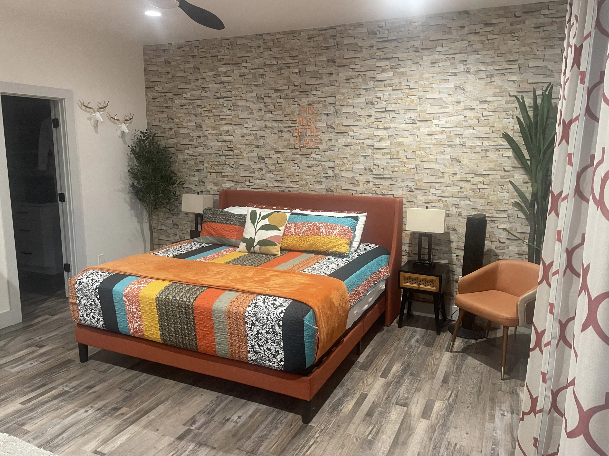 A bedroom with a bed that has a colorful quilt and pillows, a brown upholstered headboard, two nightstands with lamps, a tall plant on each side of the bed, a textured stone wall behind the bed, a small orange chair, a patterned curtain, and a ceiling fan.