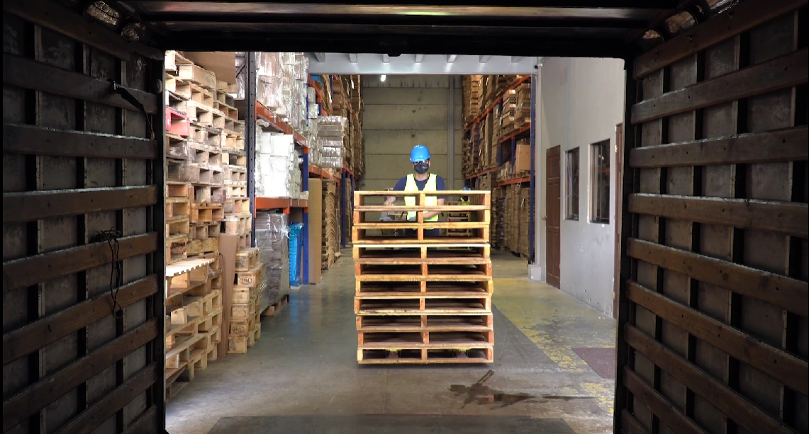 Worker in safety gear standing behind stacked wooden pallets in a warehouse aisle filled with shelves of goods.