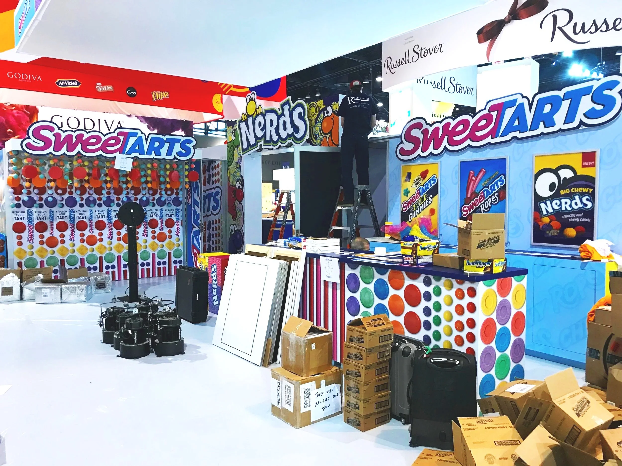 Decorated booth setup at an event, featuring colorful signage for SweetTARTS and Nerds, with boxes, lighting equipment, and construction materials in the foreground, and a person working on the booth.