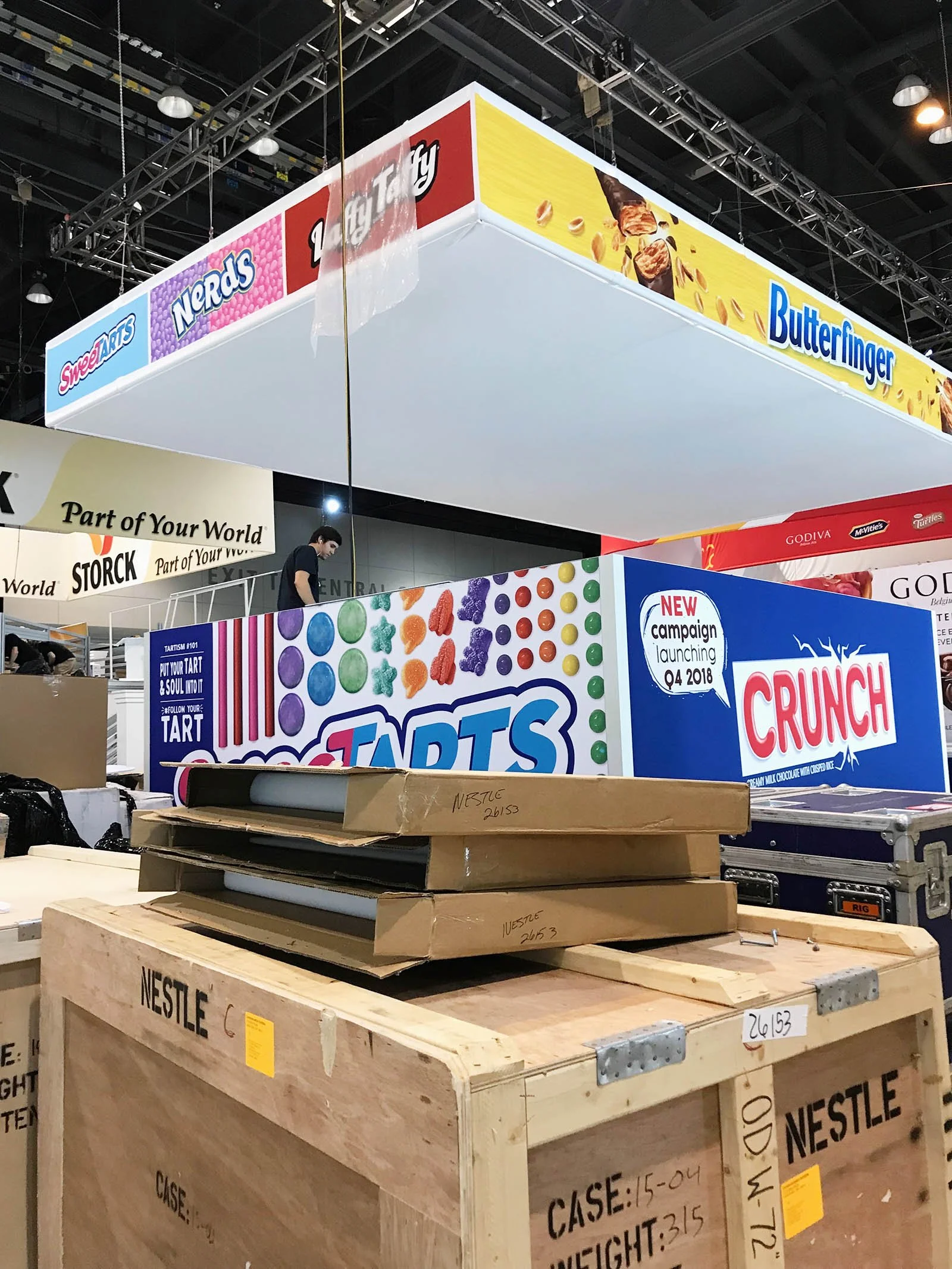 Booth setup at a trade show with large colorful signage for candy brands Tartts and Crunch, and various boxes and crates in the foreground.