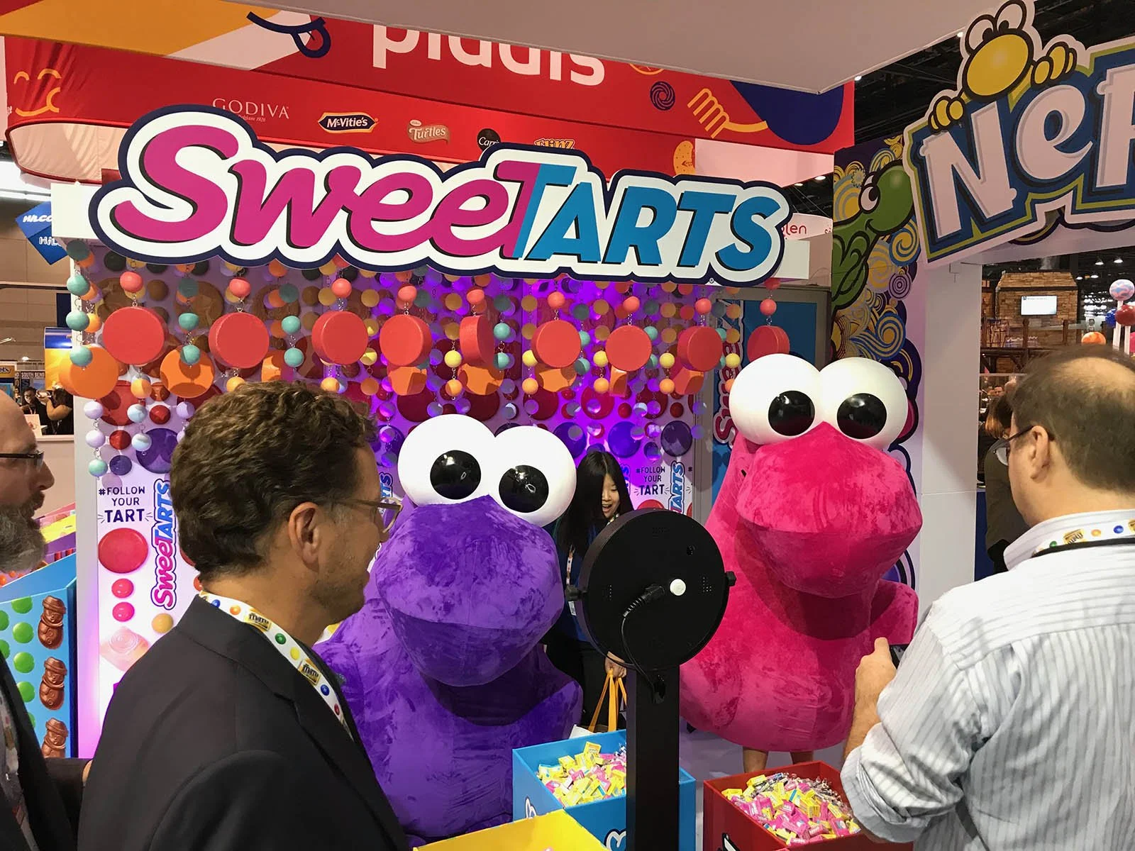 People attending a booth featuring giant purple and pink plush toys with large eyes and a sign that says SweetARTS.