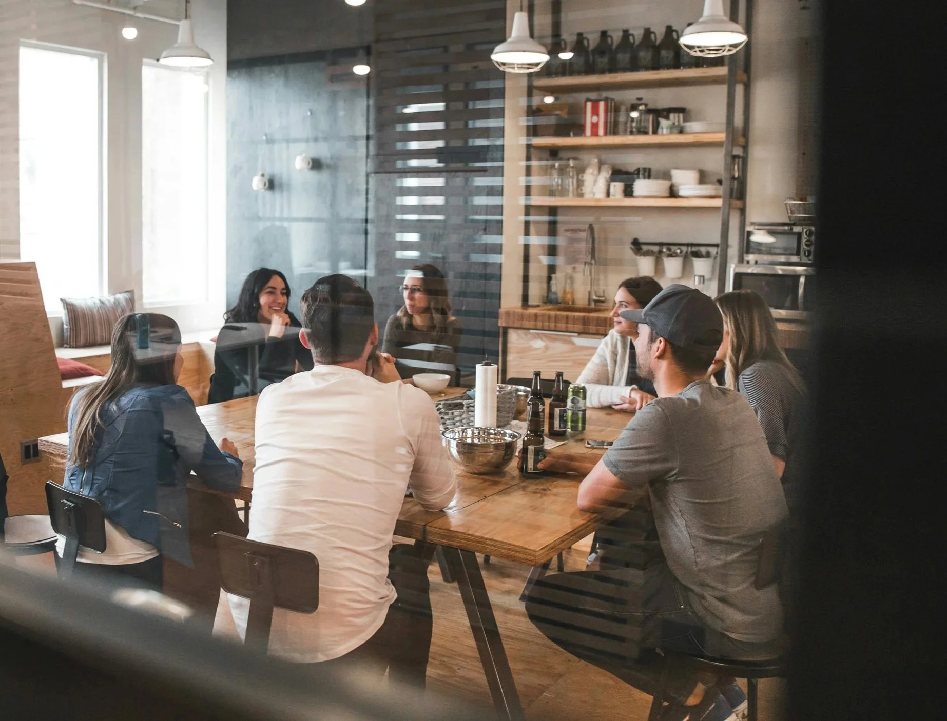 A cross-functional team sitting around a wooden dining table in a modern office kitchen, engaged in conversation and partition and open shelving in the background.
