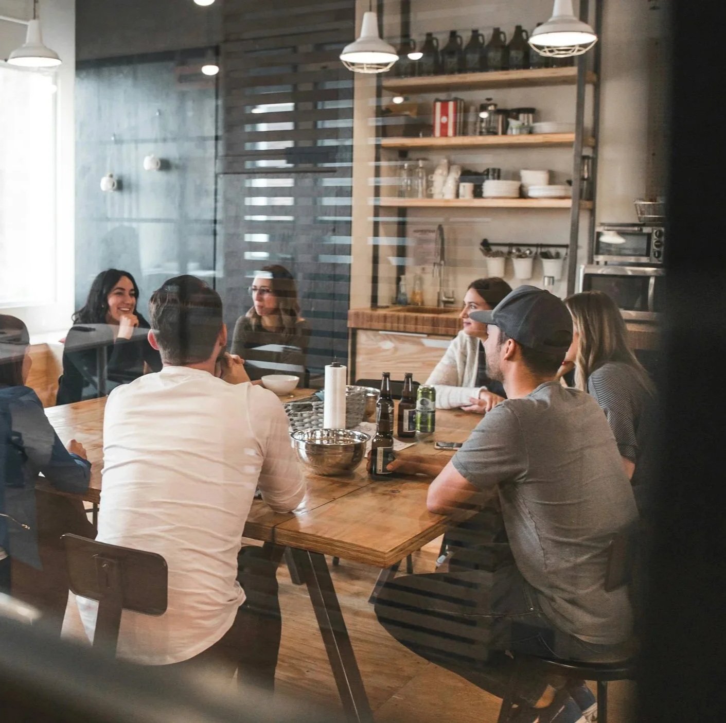 A group of six people sitting around a wooden table in a modern, cozy cafe, seen through a glass window.