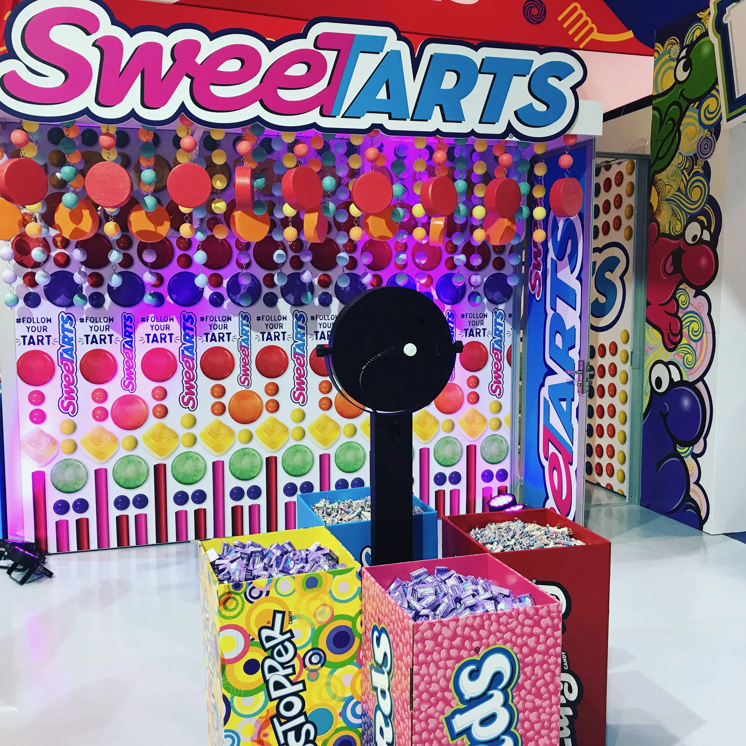 Colorful carnival-style candy toss game called Sweet Tarts with large containers of assorted candies and a black pie thrower in front of a vibrant backdrop with oversized candy graphics and signage.