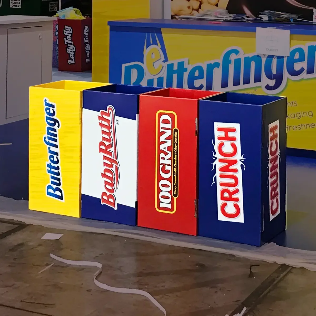 Three colorful cardboard boxes labeled Butterfinger, Baby Ruth, and Snickers, arranged in a row on a table at a candy store or display.