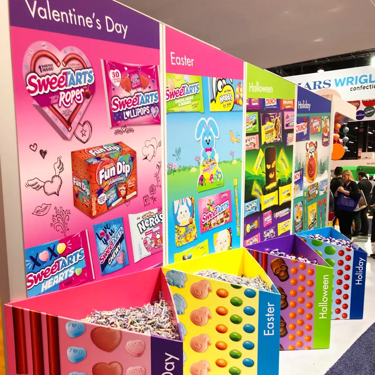 Colorful display of Valentine's Day, Easter, Halloween, and holiday candies and chocolates at a trade show or store.