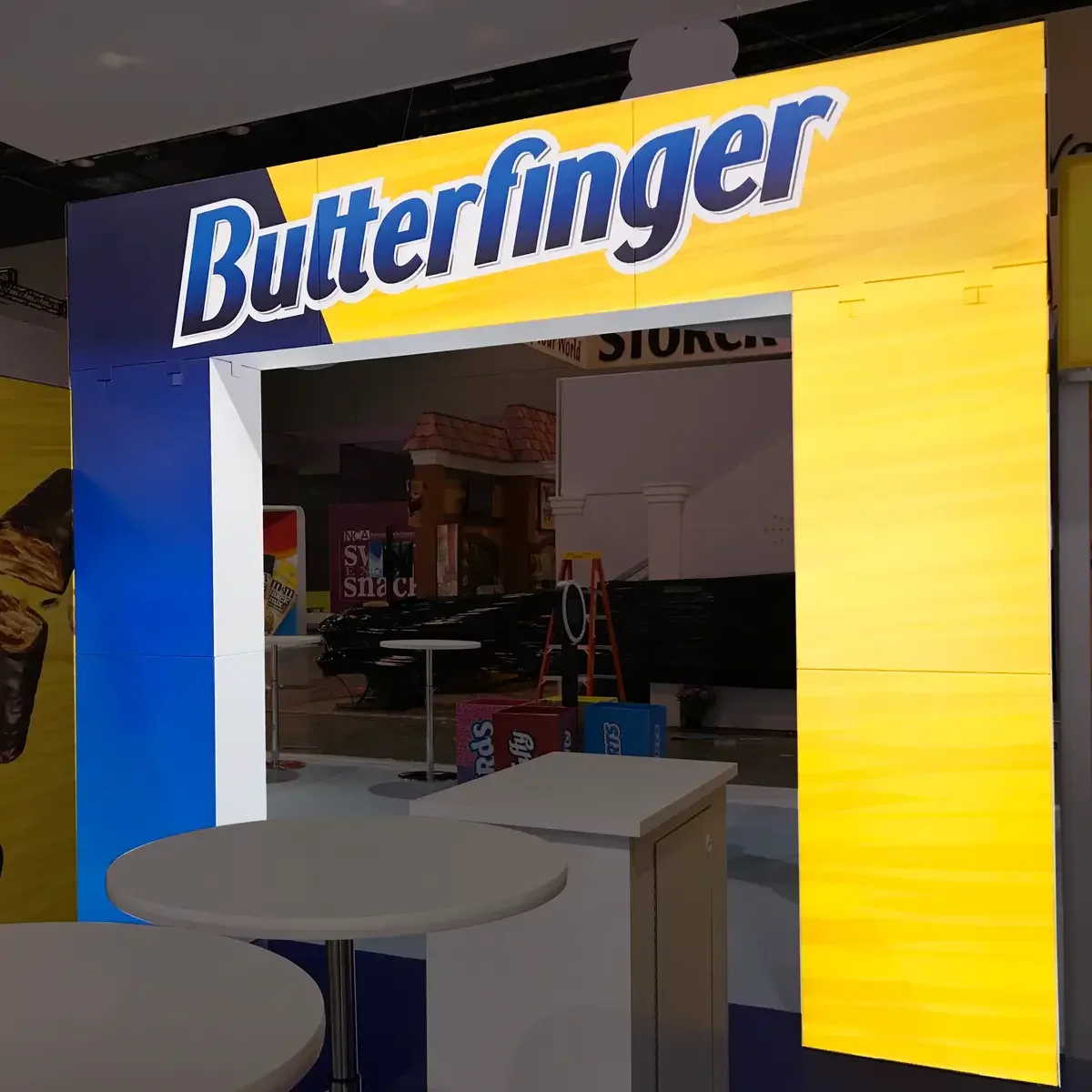 A large illuminated sign with the word 'Butterfinger' in bold blue and white letters on a yellow and blue background, hanging above an empty display booth at what appears to be a trade show or store.
