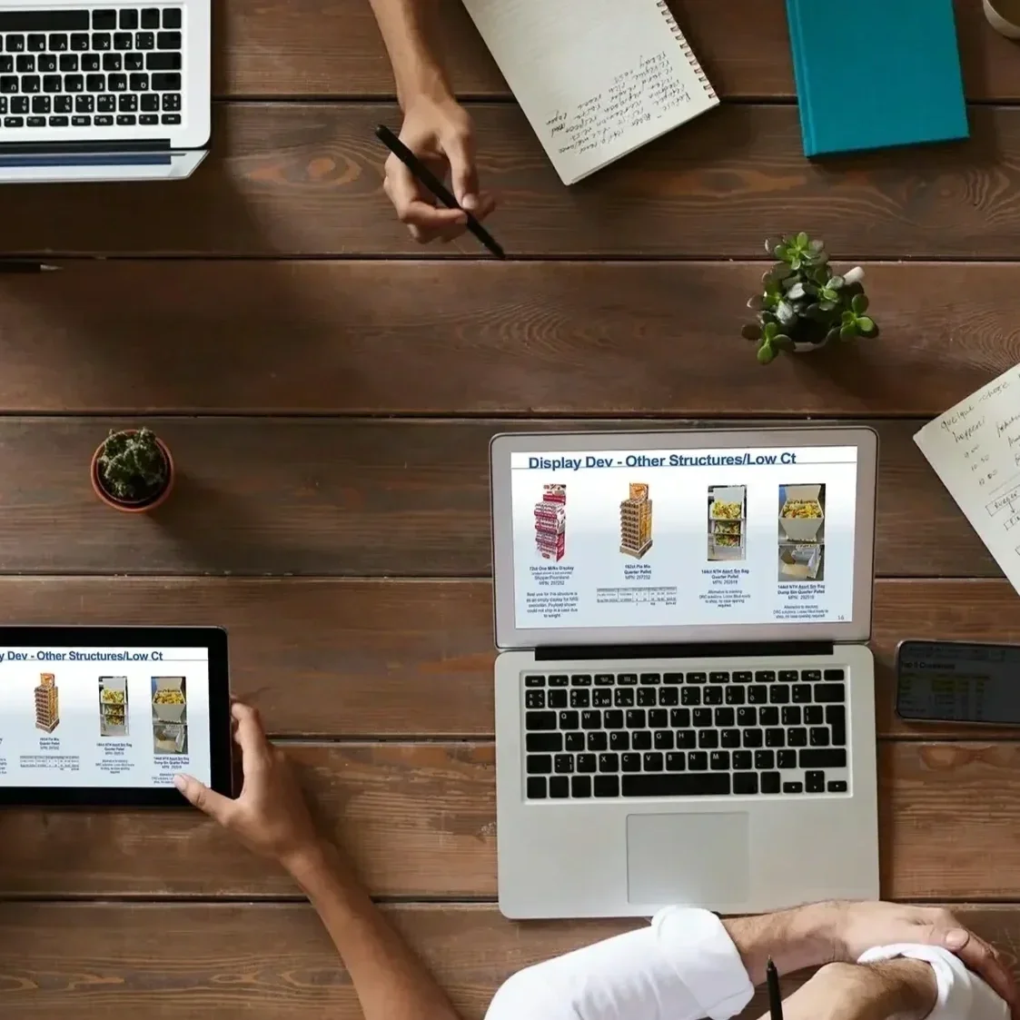 A top-down view of a wooden desk with two laptops, a tablet, a notebook, a smartphone, and two potted plants. One laptop shows a presentation slide titled 'Display Dev - Other Structures/Low Ct' with images of retail displays. The other devices display similar content. A person is holding a pen in one hand and writing in the notebook.