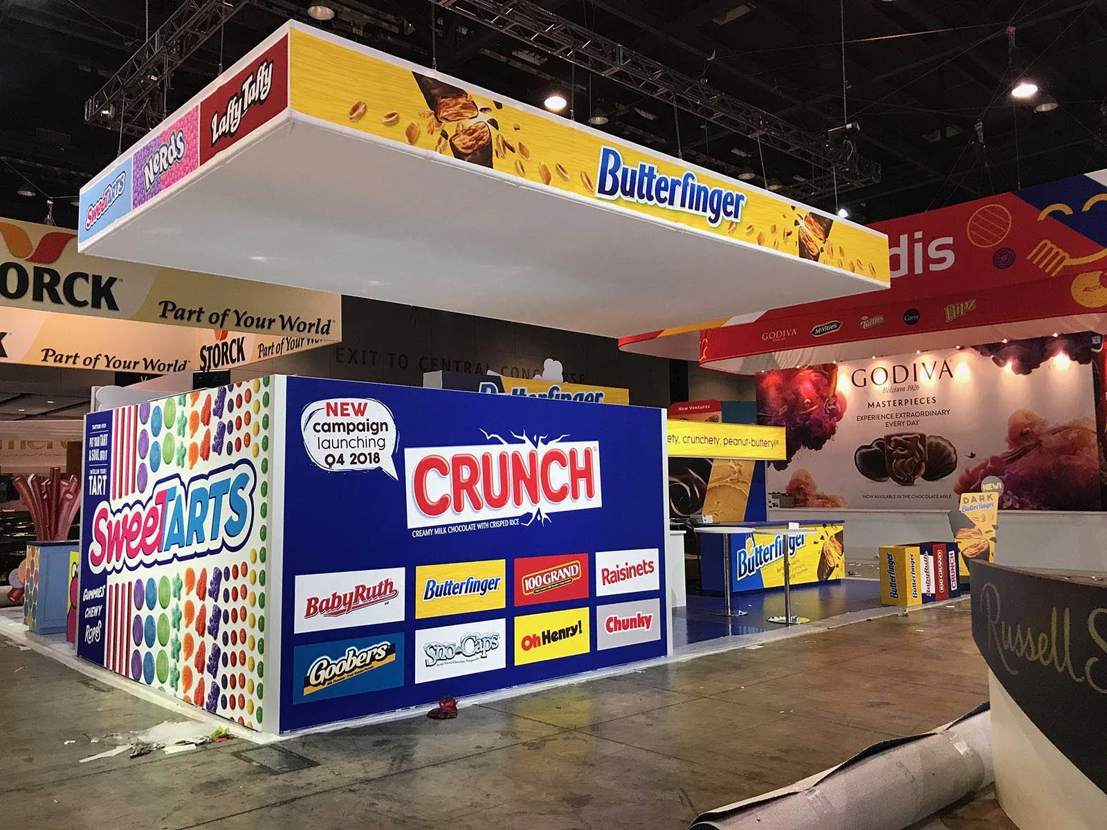 A trade show booth with branding for SweetARTS Crunch candy and Butterfinger chocolate bars, along with other snack brands, in an exhibition hall.