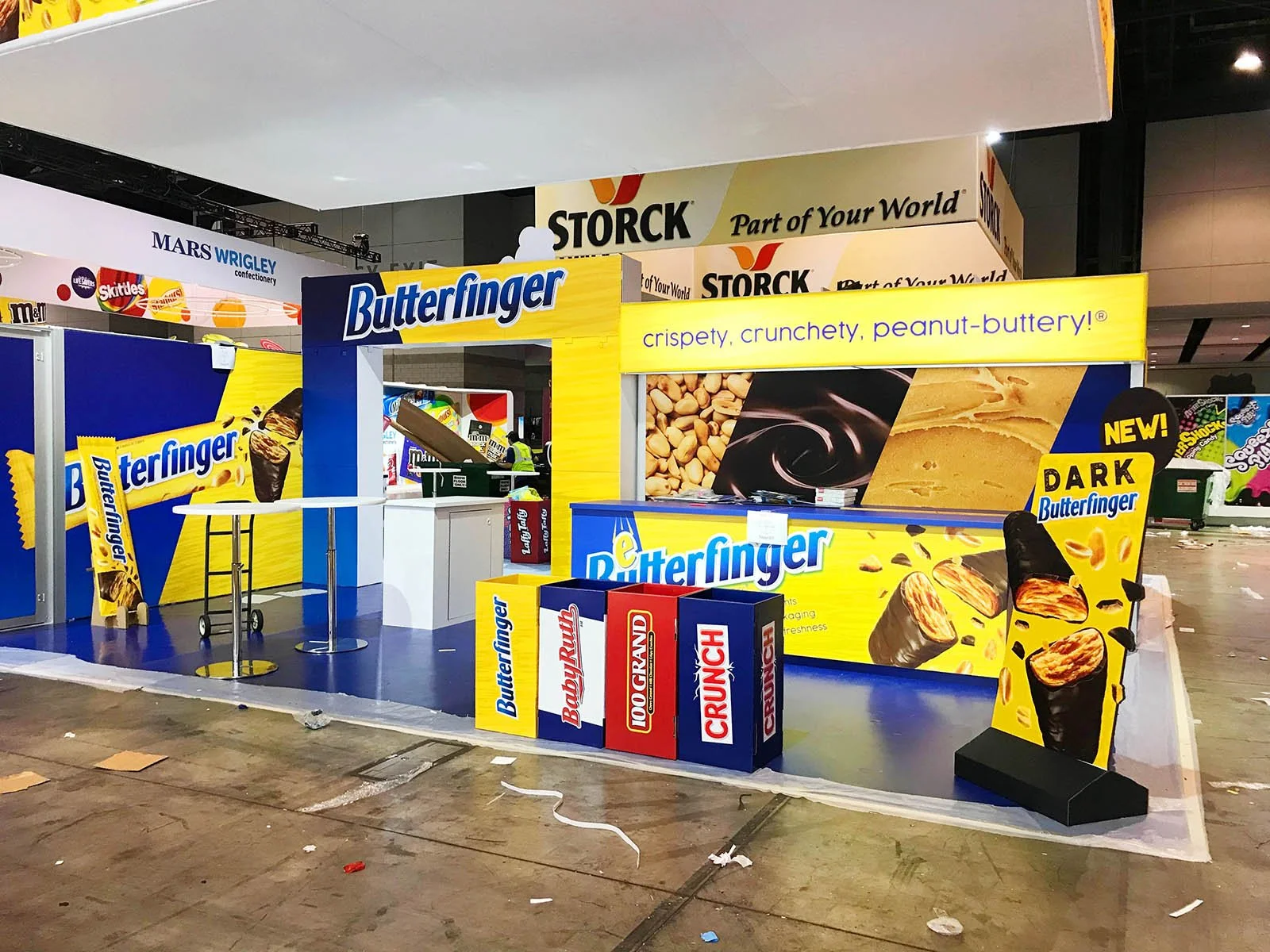 Booth display for Butterflyer peanut butter products, featuring a large yellow banner with the slogan 'crispy, crunchy, peanut-buttery!' and an image of peanut butter with peanuts, with boxes of Butterfinger, Baby Ruth, 100 Grand, and Crunch bars in 