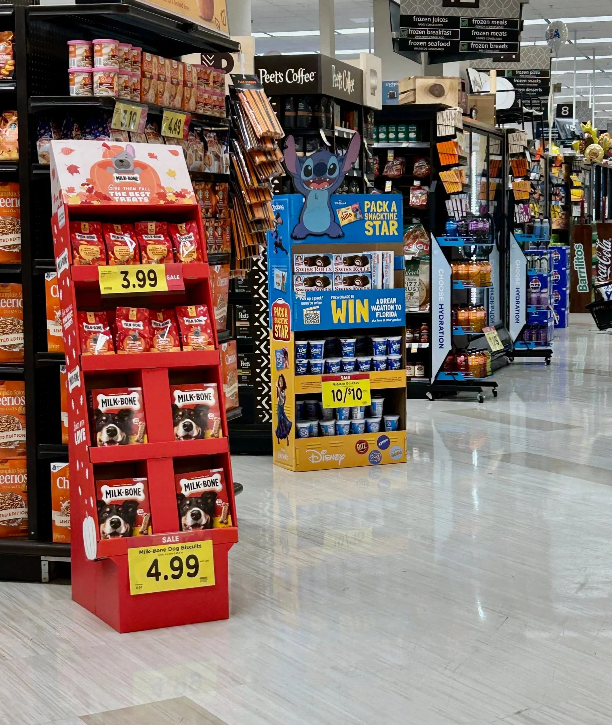 A grocery store aisle with snack displays, including a red Milk Bone dog biscuit stand and a blue Stitch plush stand promoting a prize contest, with shelves of various products and signage overhead.