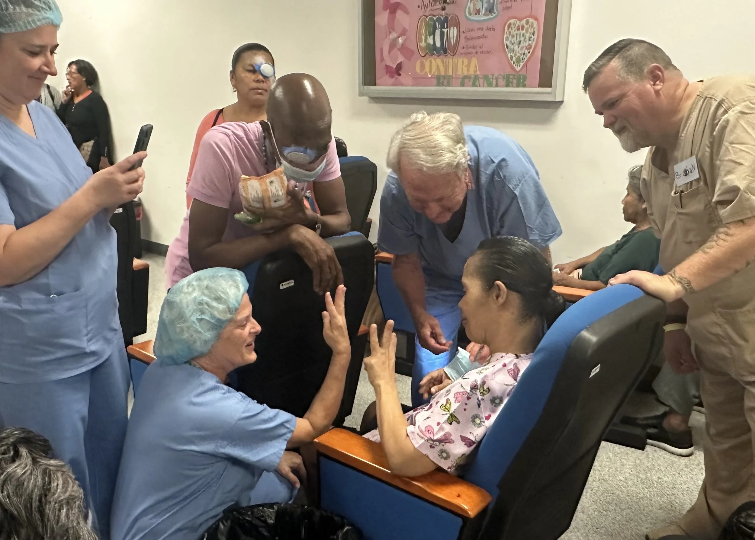 A group of medical staff and elderly patients in a healthcare setting. Two women are seated, engaging in conversation; one of the women is wearing a surgical cap and scrubs, and the other is wearing a hair cap with a hospital gown. Three other medical professionals stand nearby, one with an eye patch and mask, another leaning over the seated patients, and a third holding a smartphone. The background features a pink poster on the wall with decorative text and illustrations.