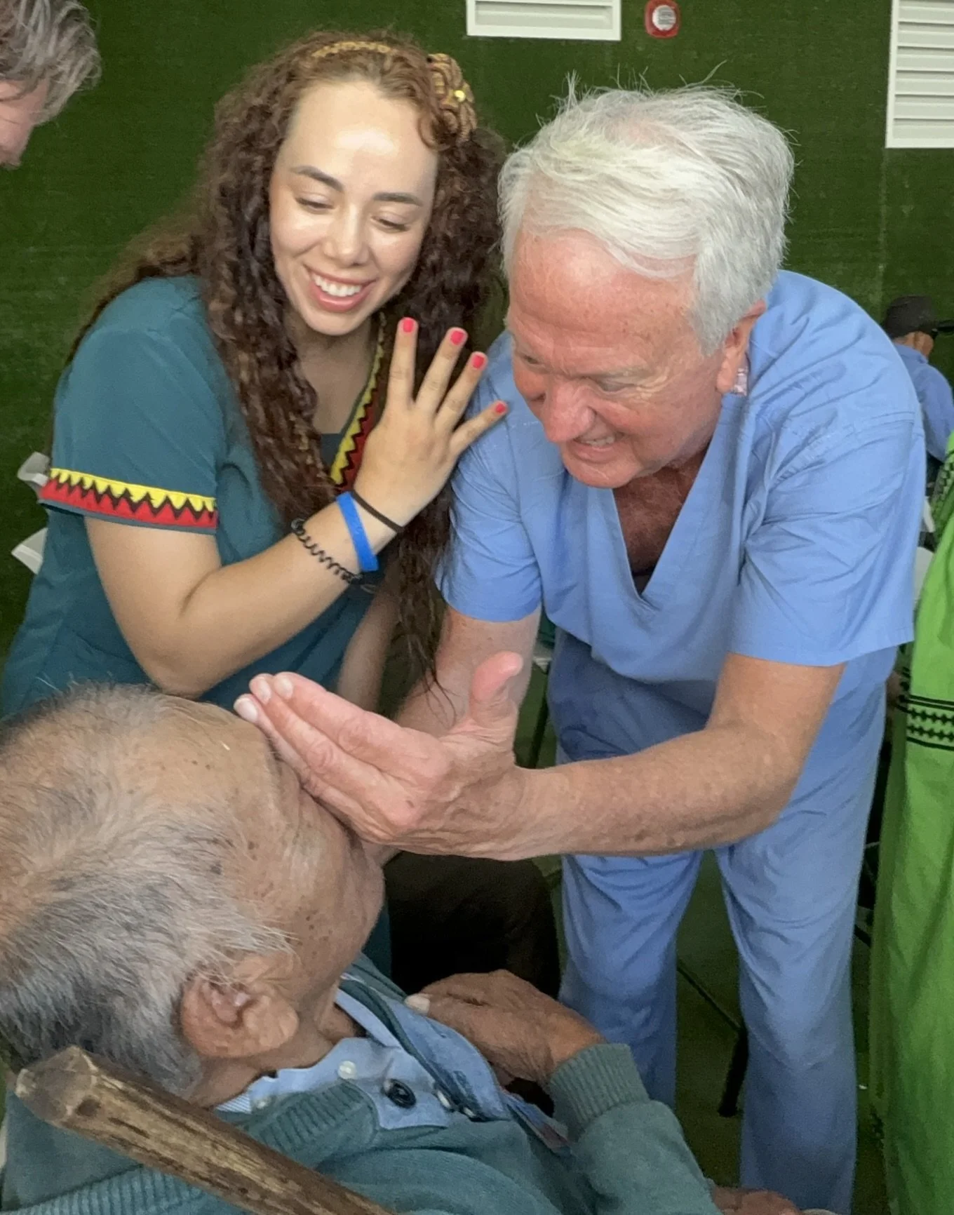 A woman with long curly hair and a blue shirt is smiling and touching the shoulder of an elderly man with white hair, wearing a blue shirt, who is smiling back. They are interacting in a green indoor setting.