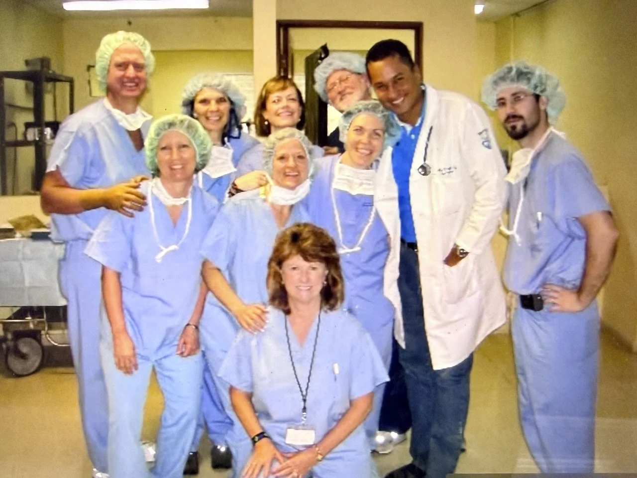 Group of healthcare professionals in scrubs and hair covers, smiling in a hospital setting.