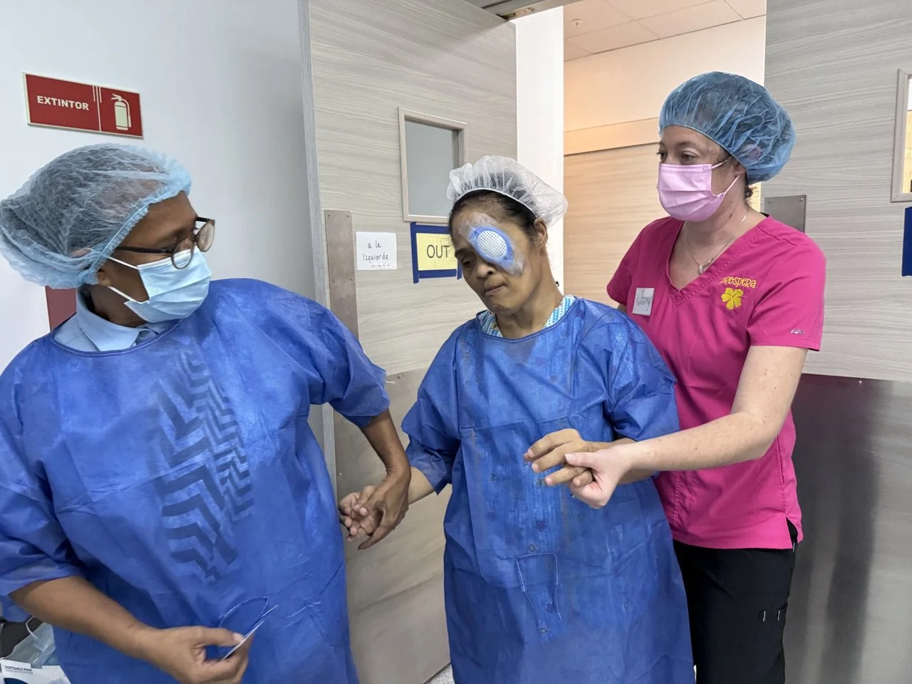 Three healthcare workers in PPE, including masks, hair covers, and gowns, are attending to a patient with a facial injury in a hospital corridor.