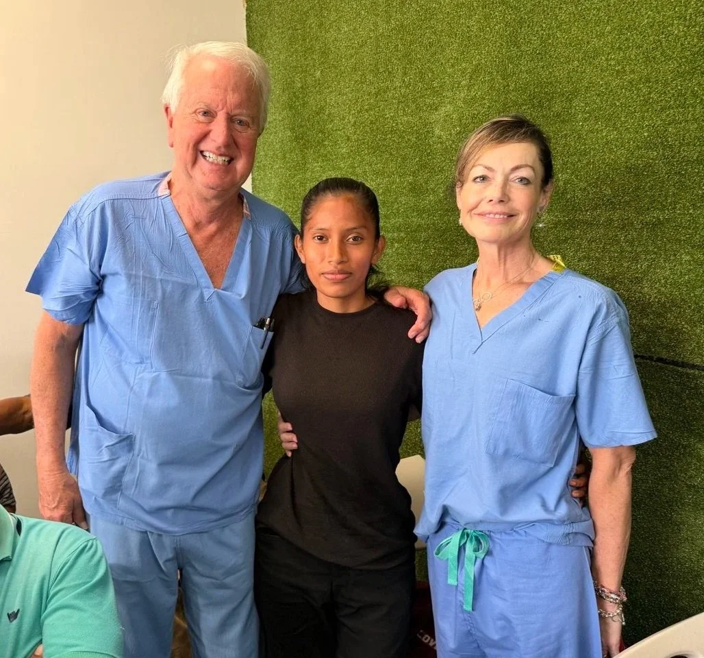 Three medical professionals, two women and one young woman, smiling and standing closely together in a medical setting. The men and women are wearing scrubs and are in front of a green wall.