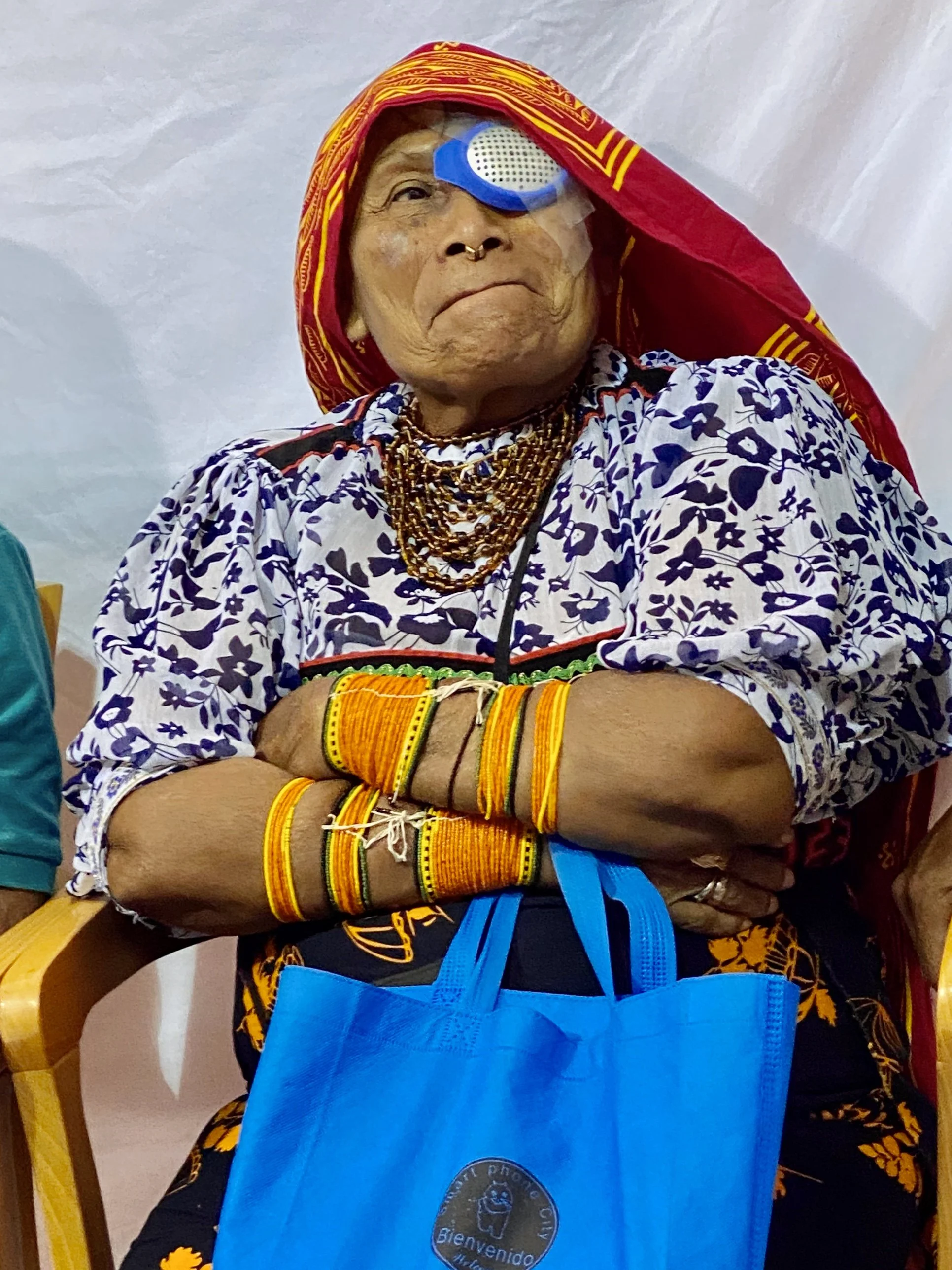 Elderly person with traditional tribal jewelry and clothing, wearing a red headscarf, a blue plastic eye patch, and a blue apron with a logo that says "Bienvenido".