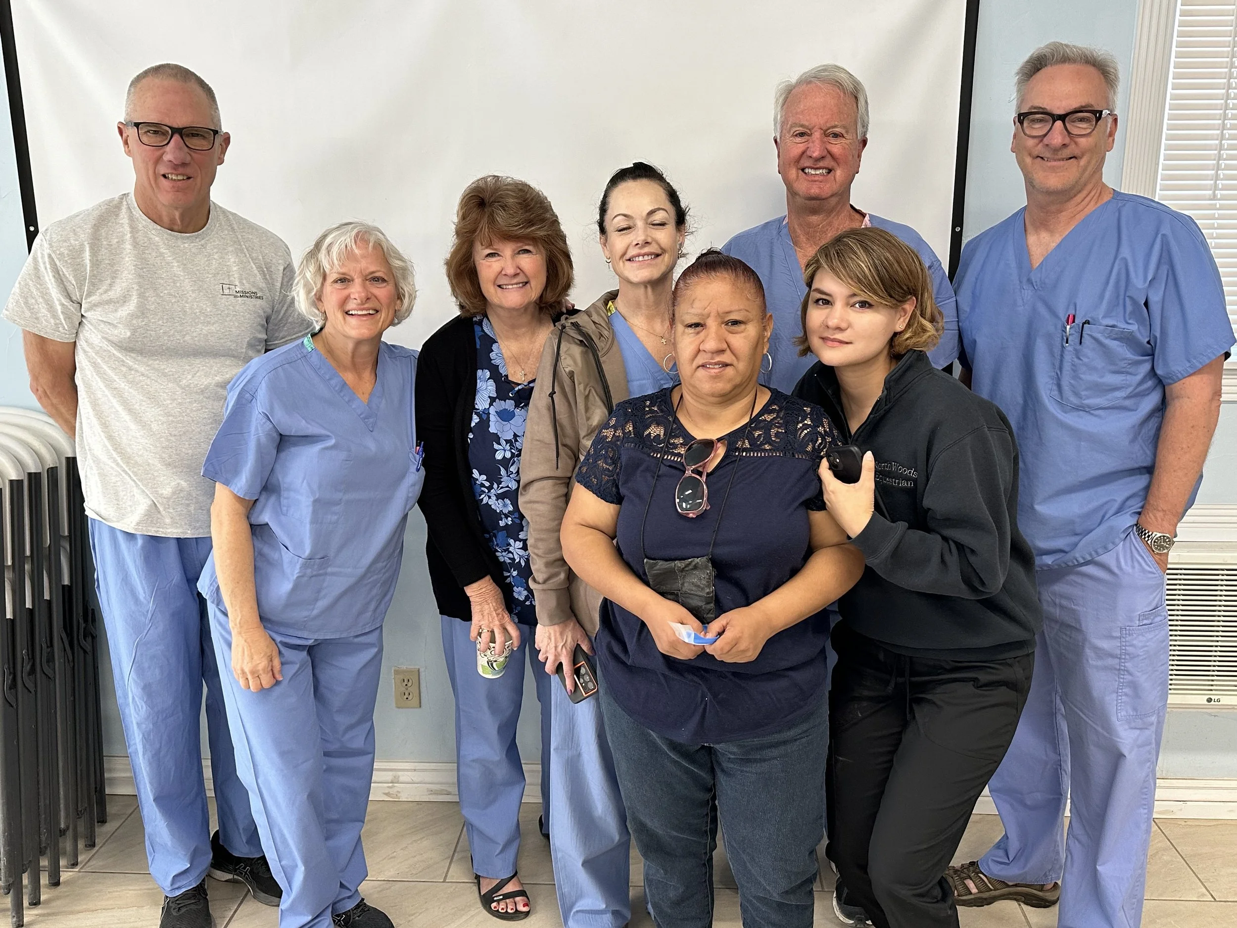 Group of nine medical professionals and staff members standing together in a hospital or clinic, some in scrubs, smiling at the camera.