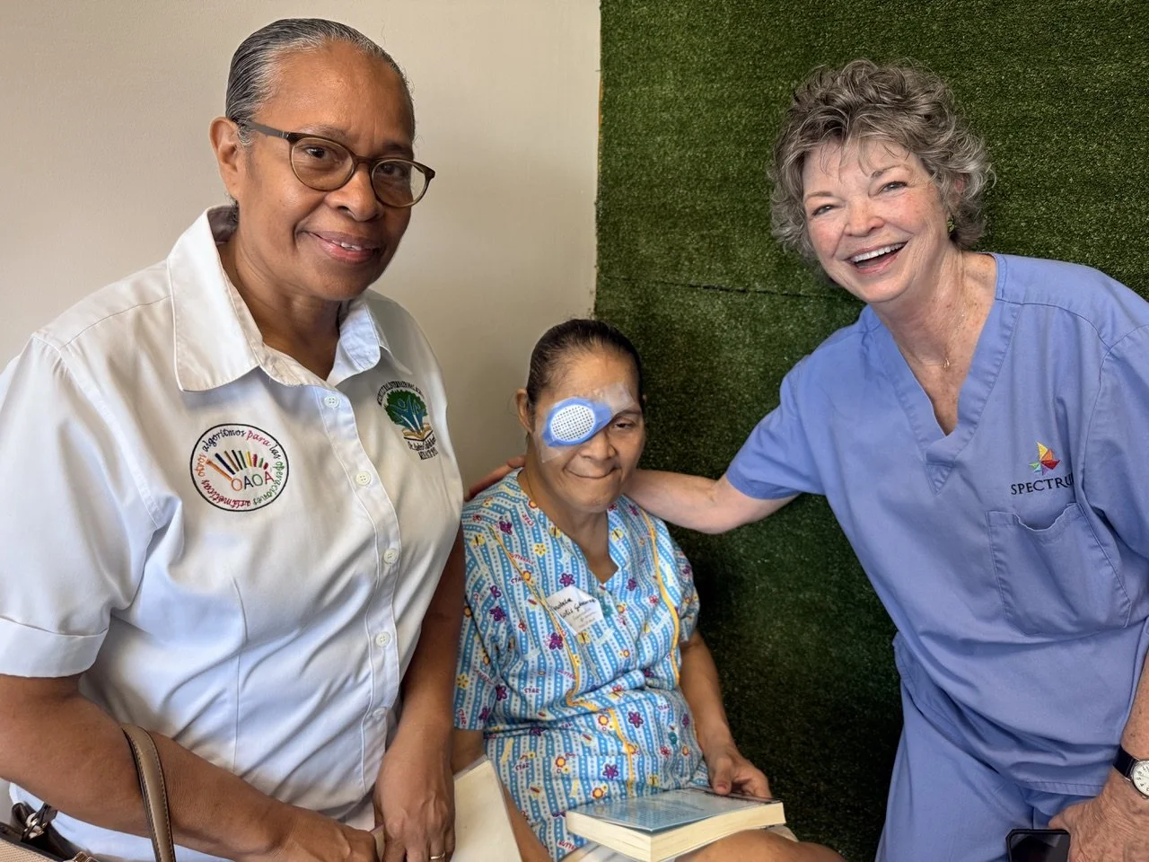 Three women, one patient with an eye patch, smiling and posing for a photo indoors. The patient is wearing hospital gown with colorful patterns. Two healthcare workers are standing on either side, one in a white shirt and the other in blue scrubs, both smiling. The woman in blue scrubs has her hand gently on the patient's shoulder.