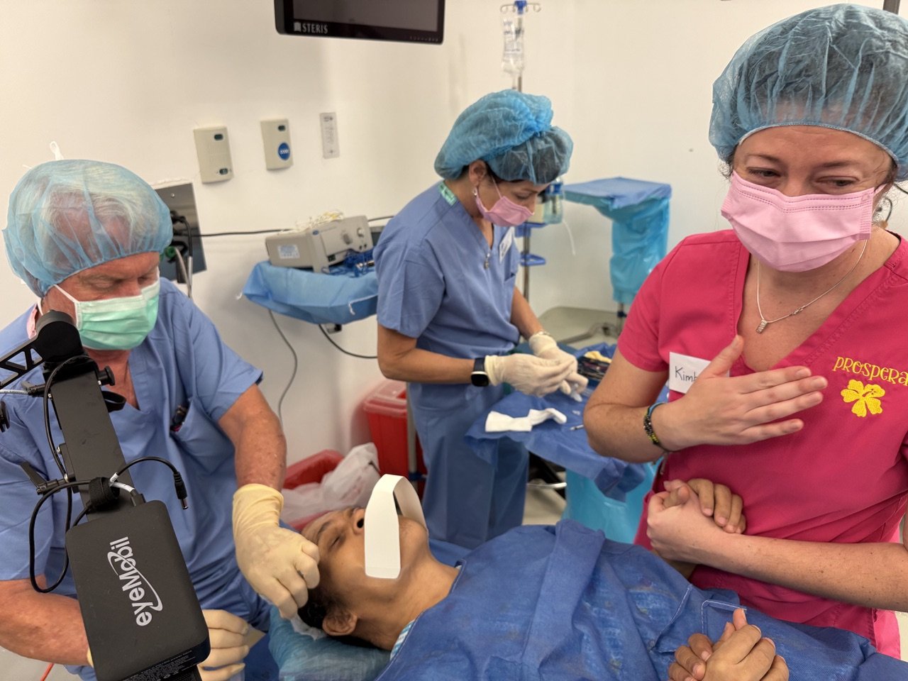 Medical team performing surgery on a patient in an operating room, with healthcare professionals wearing scrubs, masks, and gloves, and one person doing a hand heart gesture.