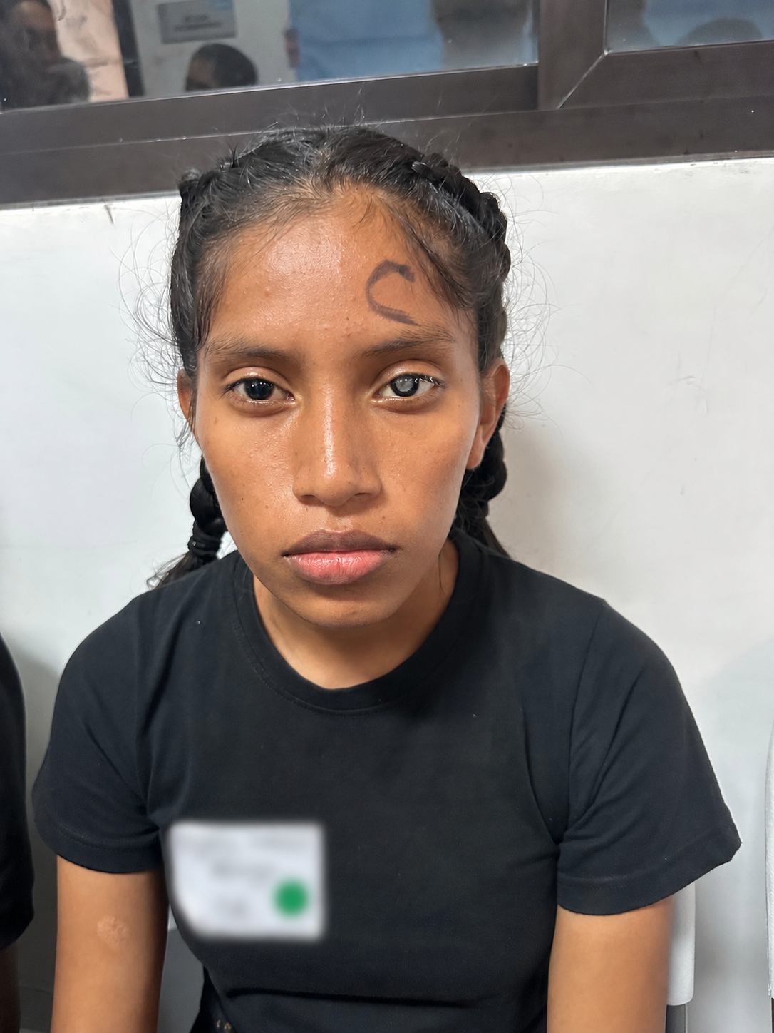 A young woman with braided hair and light skin, sitting against a white wall with a mirror behind her, wearing a black t-shirt, and looking directly at the camera.