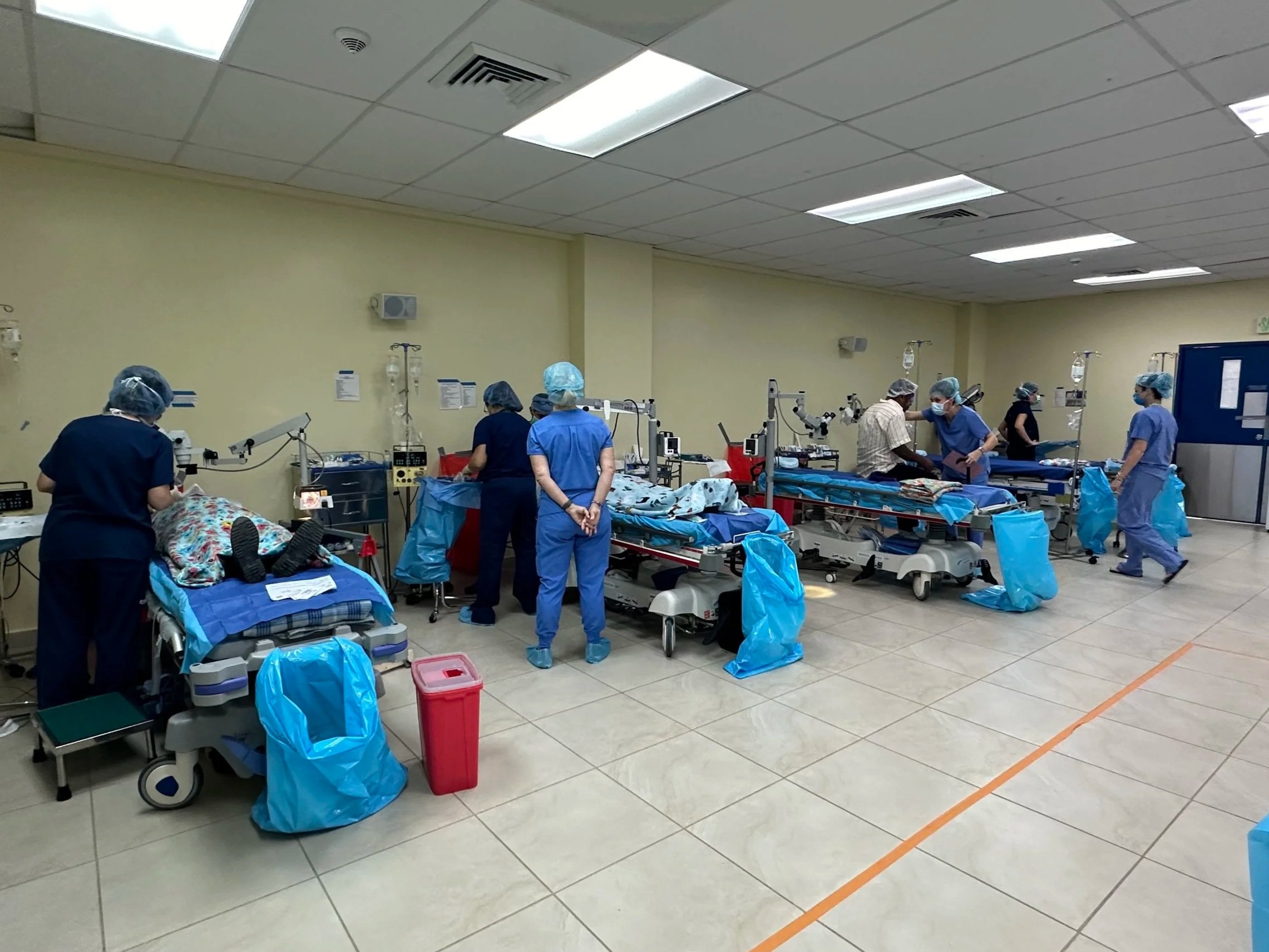 Hospital operating room with medical staff and equipment, including beds, IV stands, and monitors, with a beige wall and ceiling lights.