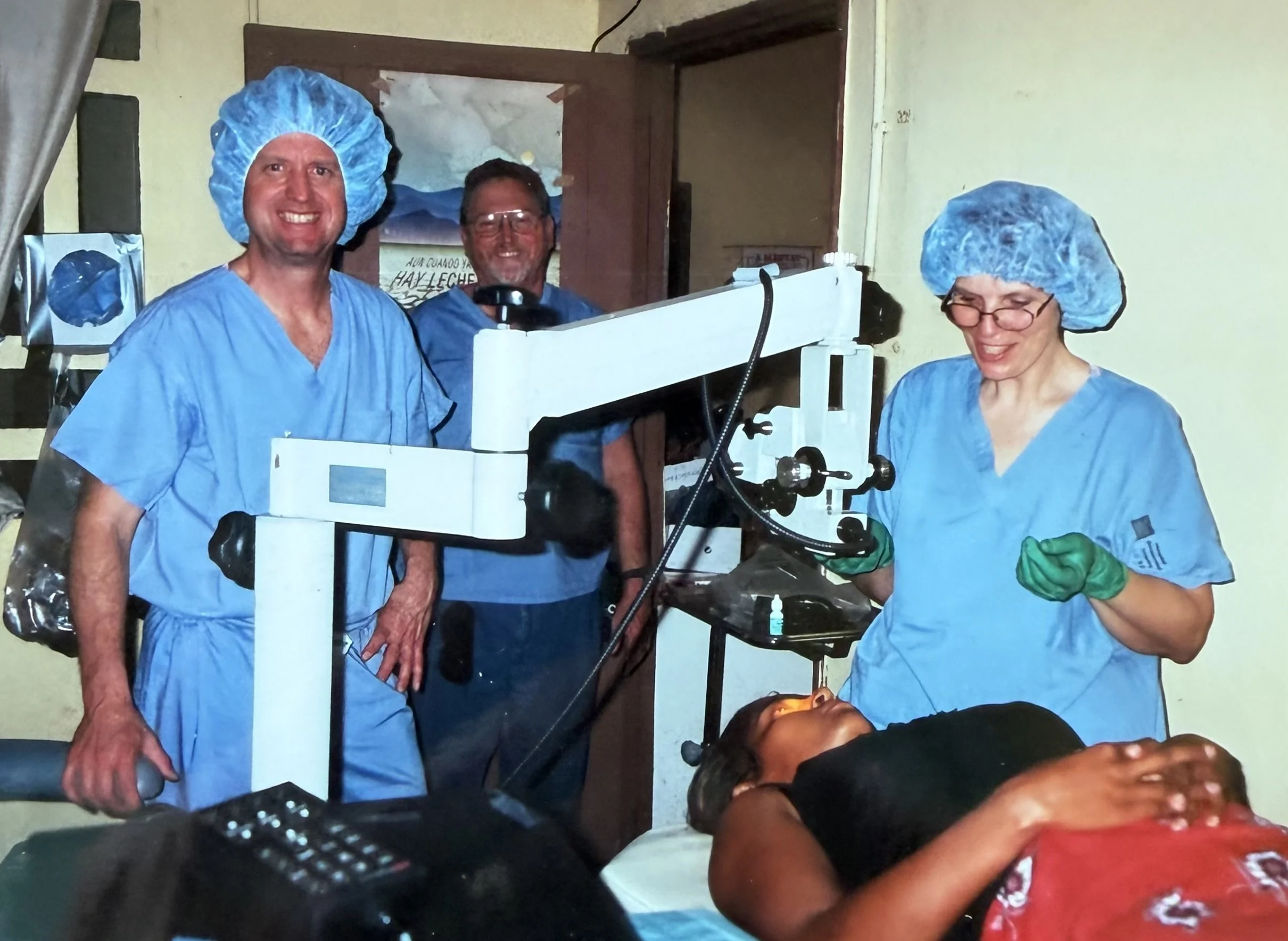 Three medical professionals dressed in blue scrubs, hair caps, and gloves, working together in a clinical setting. One is smiling at the camera, while a woman patient is lying down in front of them with her eyes closed.