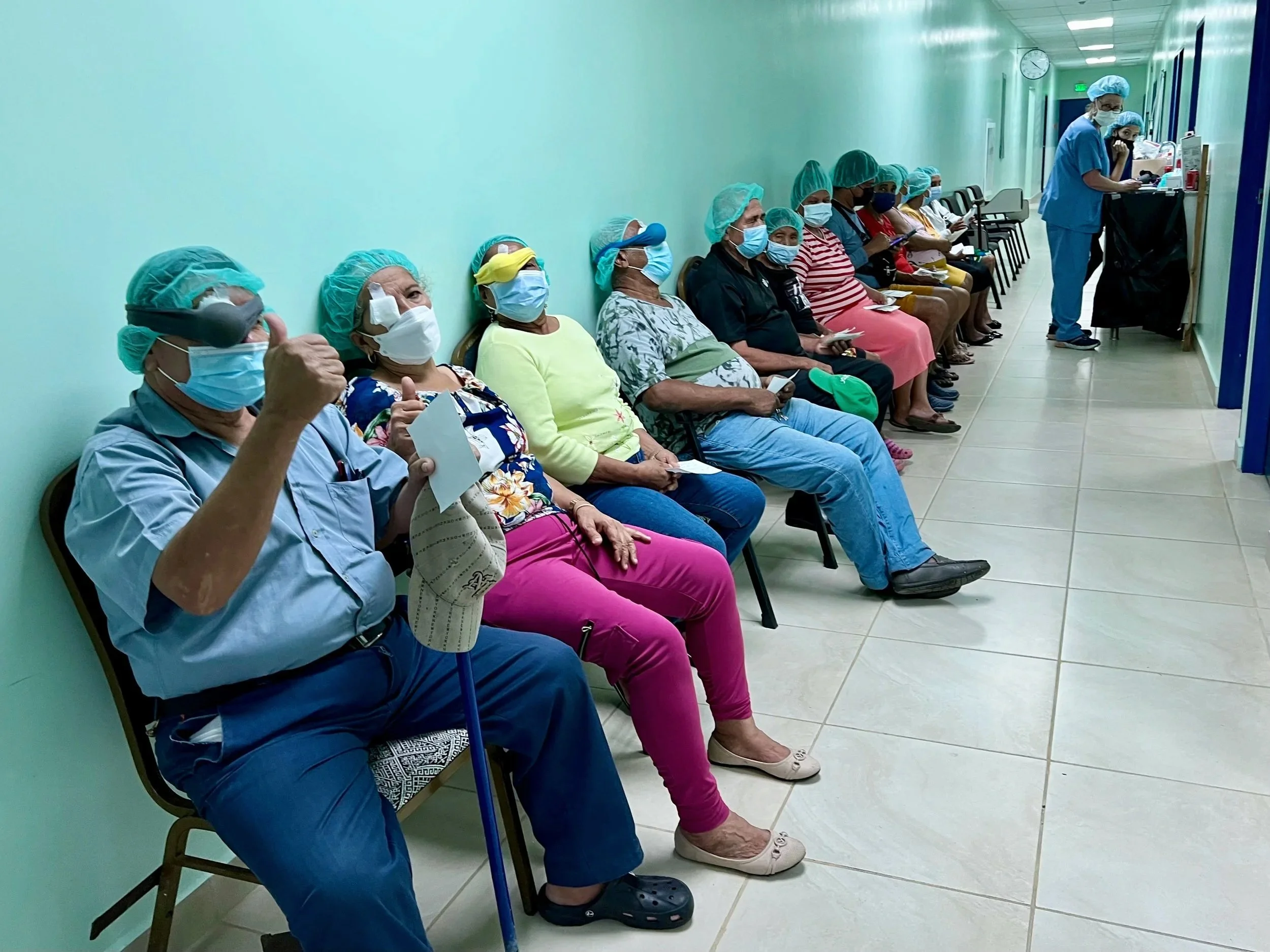 People sitting in a line, all wearing face masks and medical caps, at a healthcare facility hallway, possibly a vaccination or check-in area.