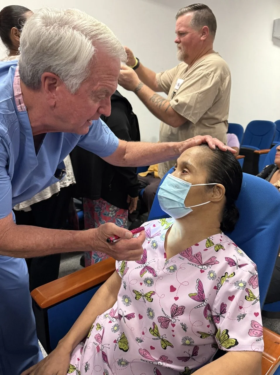 A woman in a pink floral hospital gown and face mask sitting in a medical chair, receiving a vaccination administered by a healthcare worker with white hair and blue scrubs, while another healthcare professional in beige scrubs prepares a syringe in the background.