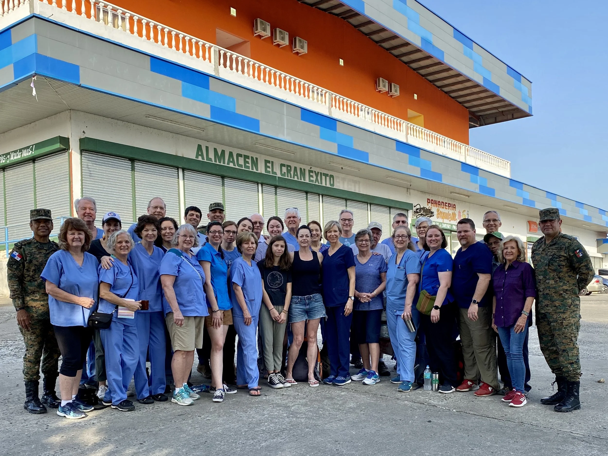 A large group of diverse people gathered outside a colorful building with an orange upper floor, blue and gray lower walls, and a sign that reads "Almacen El Gran Exito." Many are dressed in blue scrubs, suggesting they might be healthcare workers or volunteers. They are smiling and posing for a group photo.