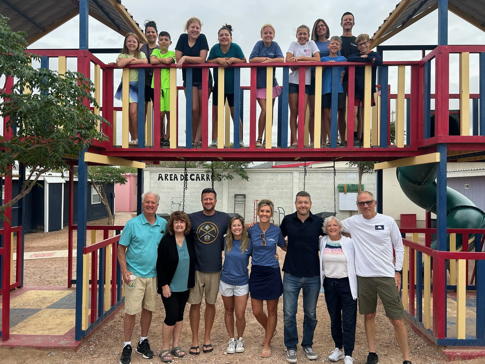 Group of 12 people, adults and children, posing under a colorful playground structure at a park on a cloudy day.