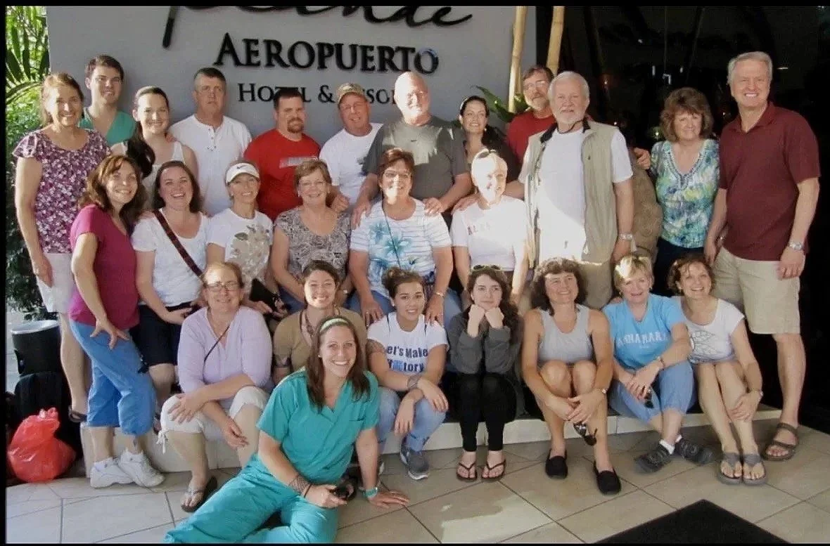A group of people standing on and sitting in front of a sign that reads 'AEROPUERTO Hotel & Resort'. The group includes both men and women of various ages, smiling and posing for the photo.