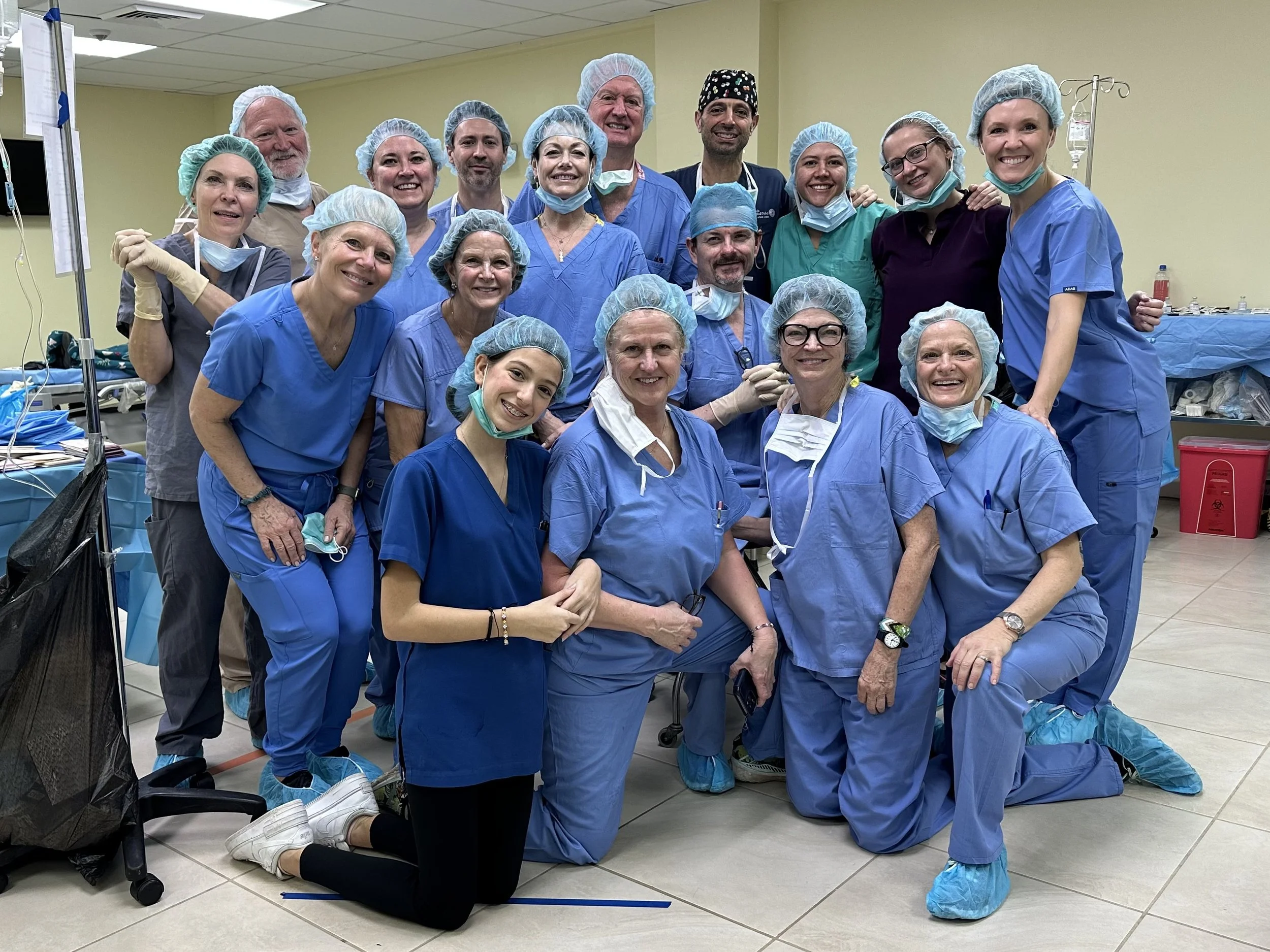 A group of healthcare professionals wearing blue scrubs, surgical caps, masks, and shoe covers posing together in a hospital or clinic setting. They are smiling and appear to be celebrating or commemorating an event.