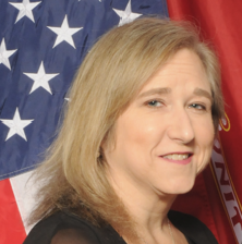 A woman with blonde hair standing in front of the American and USMC flags, smiling slightly.