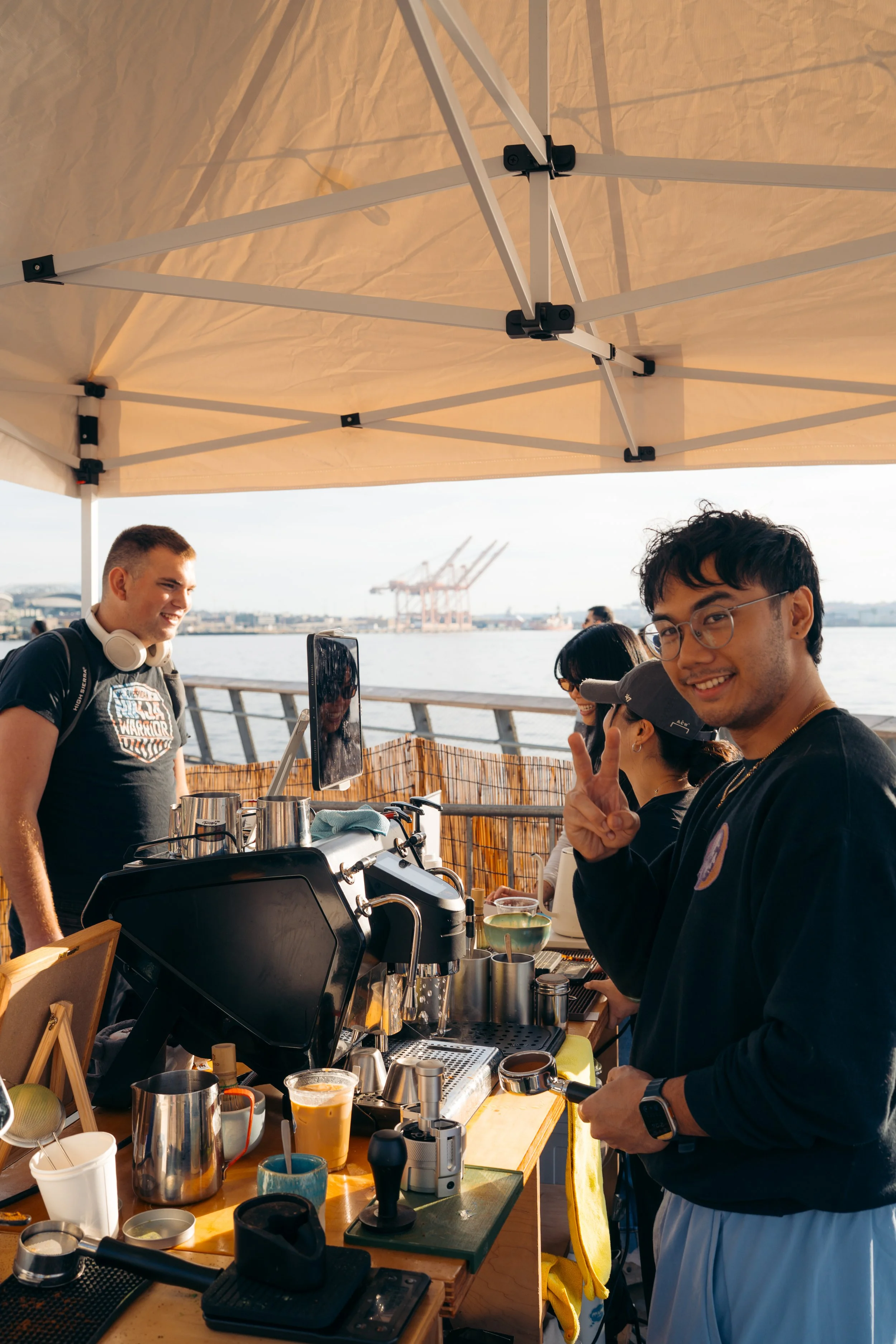 People serving and enjoying coffee at an outdoor waterfront café under a beige canopy, with shipping docks and cranes in the background.