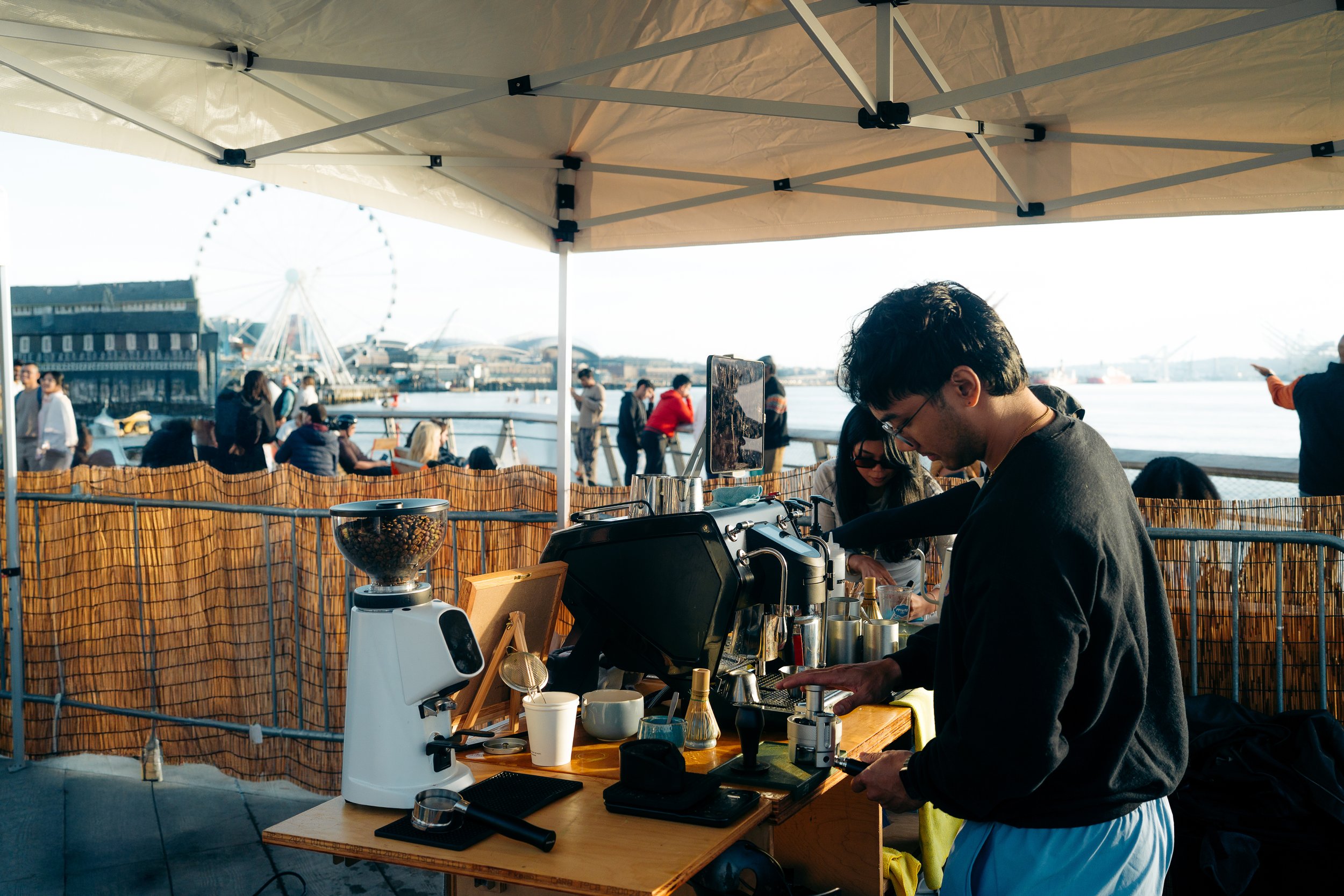 A barista making coffee at an outdoor coffee stand under a canopy with a view of a waterfront, Ferris wheel, and people walking and sitting in the background.
