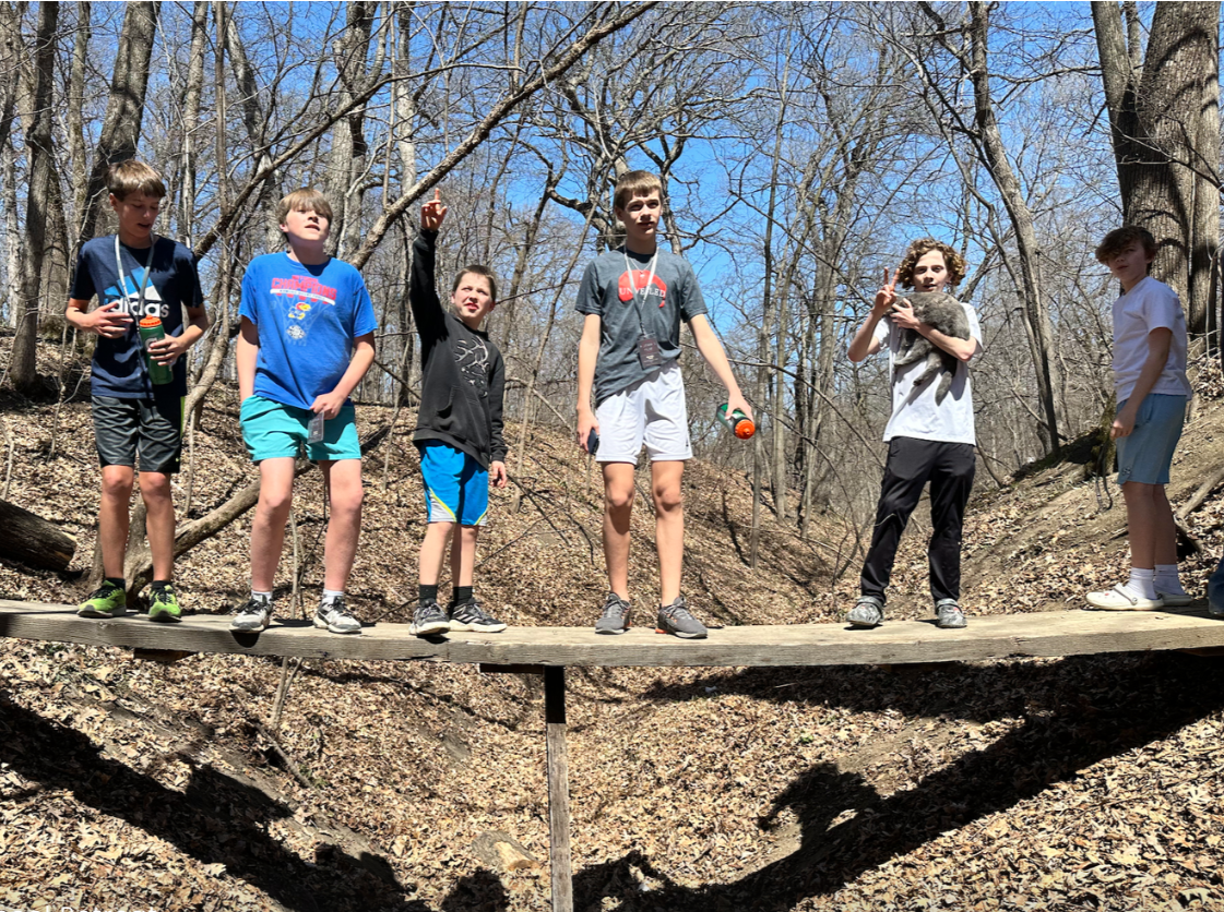 Six boys standing on a wooden bridge in a wooded area with bare trees and a blue sky.