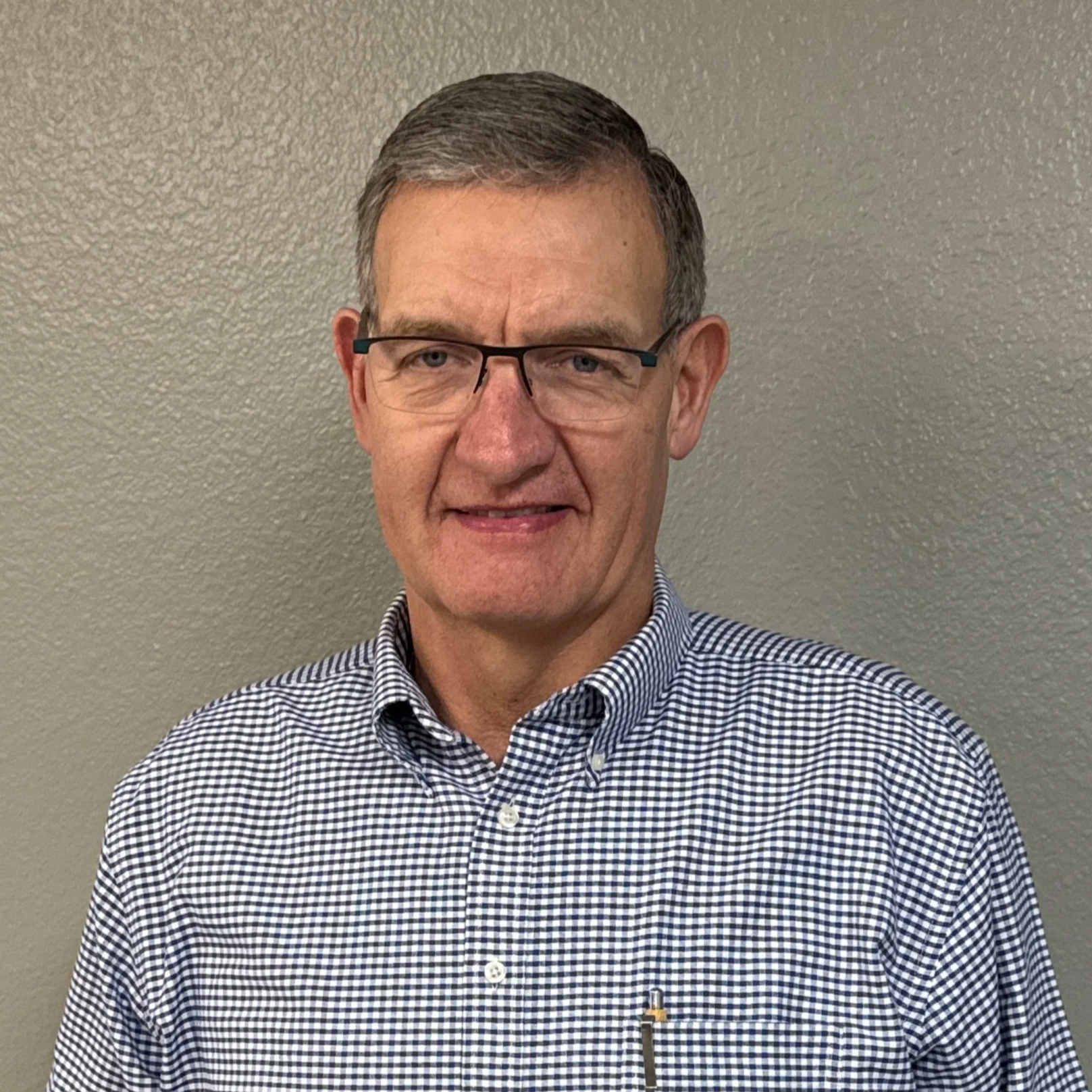 A middle-aged man with glasses, short brown hair, and a checkered shirt standing against a plain beige wall.